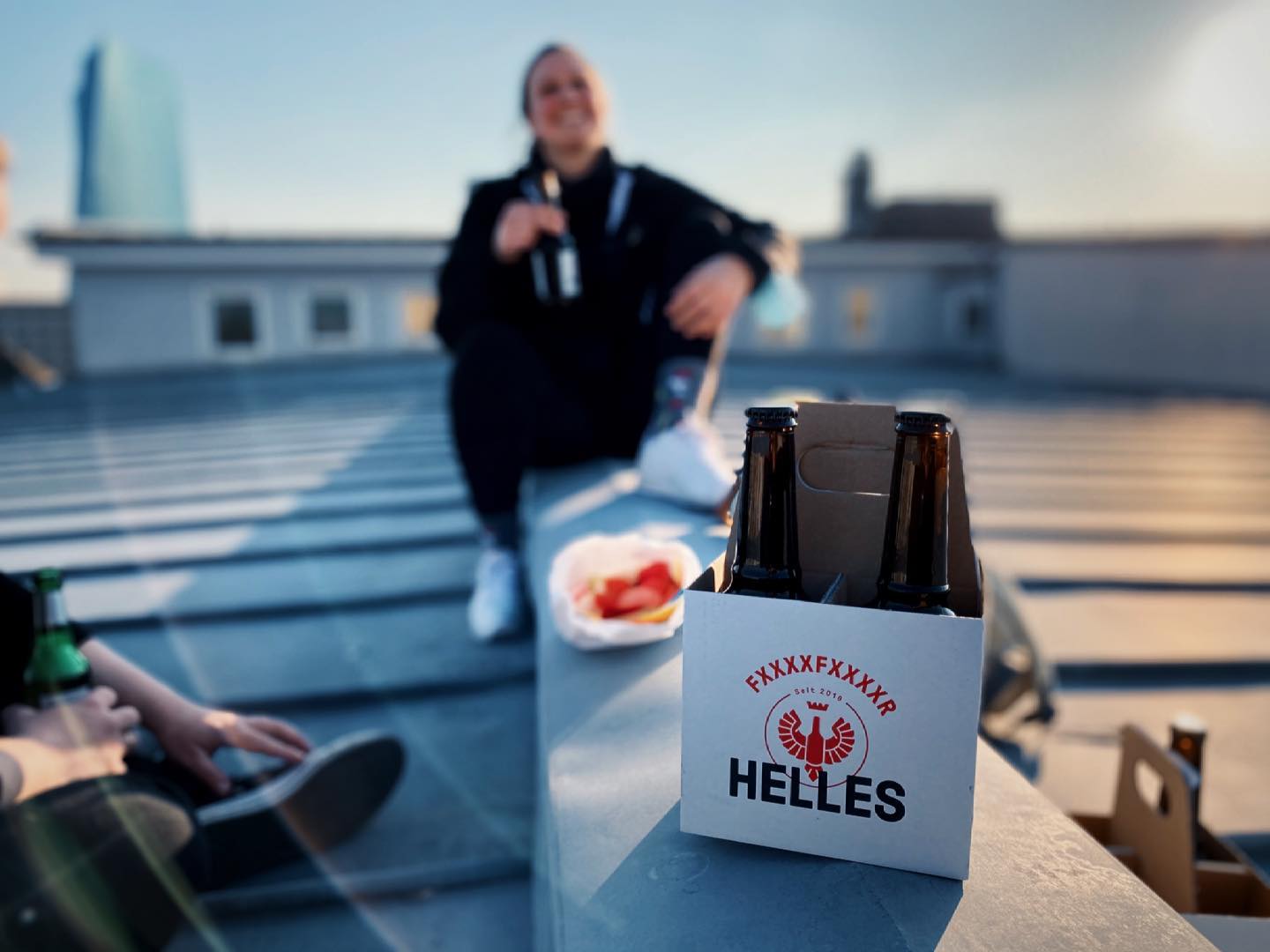 Eine Person sitzt auf einer Dachterrasse mit Getränken und Snacks, im Vordergrund ein Sechserpack Helles-Bier.