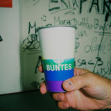 Eine Hand hält einen Magic Becher BUNTES *LIMITED EDITION*; die Wand ist mit farbenfrohen Regenbogen-Graffitis übersät.