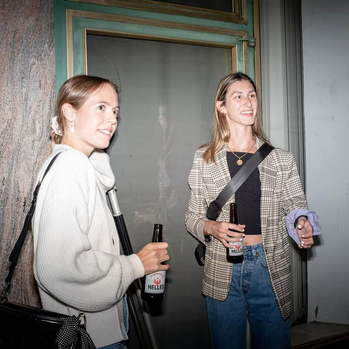 Zwei Frauen stehen lächelnd und mit Bierflaschen in der Hand in einem Haus in der Nähe eines Fensters zusammen.