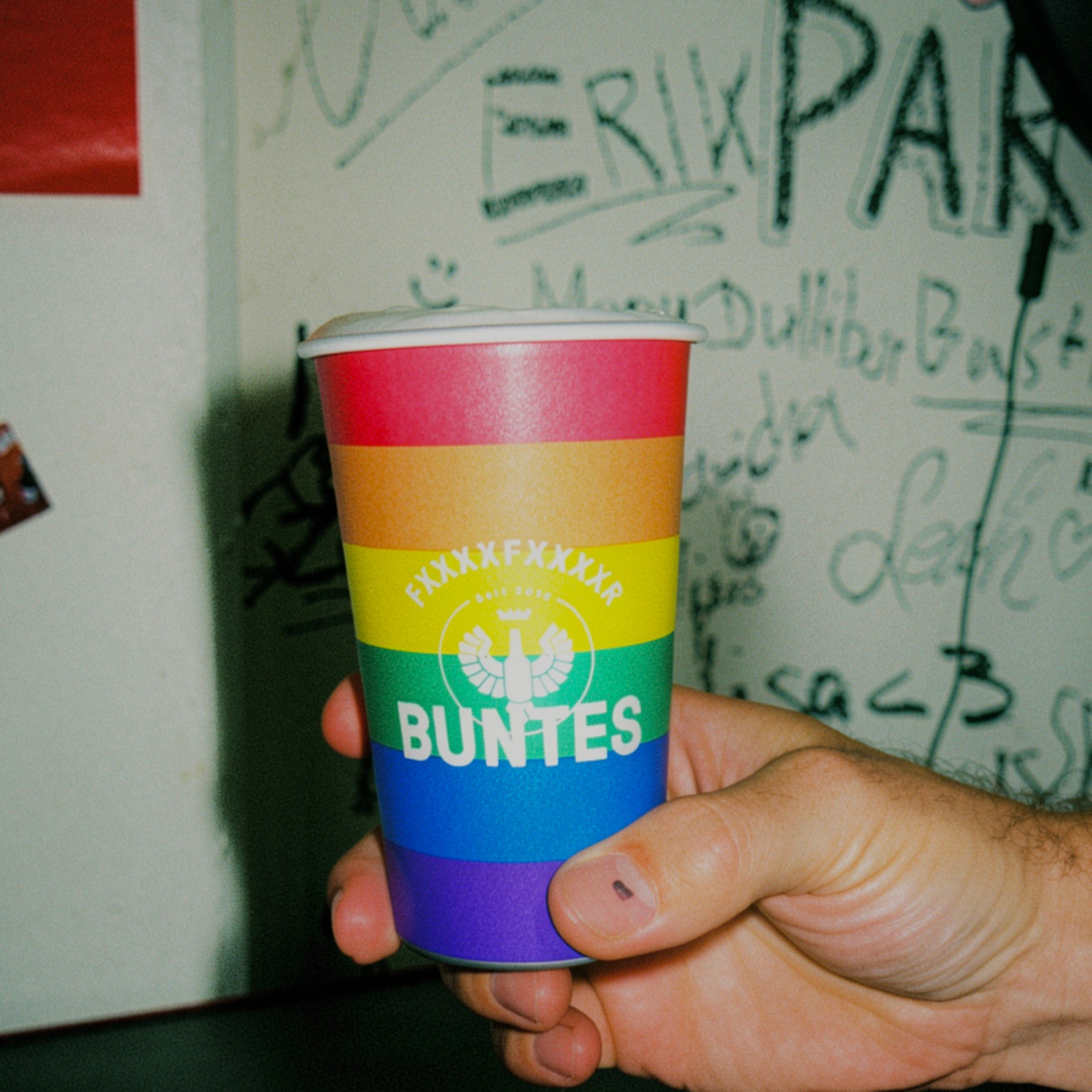 Eine Hand hält einen Magic Becher BUNTES *LIMITED EDITION* mit einem weißen Adler-Logo; im Hintergrund ist Graffiti zu sehen.