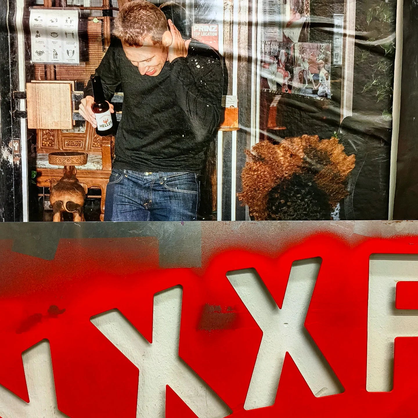 Ein Mann, der ein Bier in der Hand hält, reagiert, als Flüssigkeit auf ihn spritzt; im Vordergrund ist ein großes rotes XXX-Zeichen zu sehen.