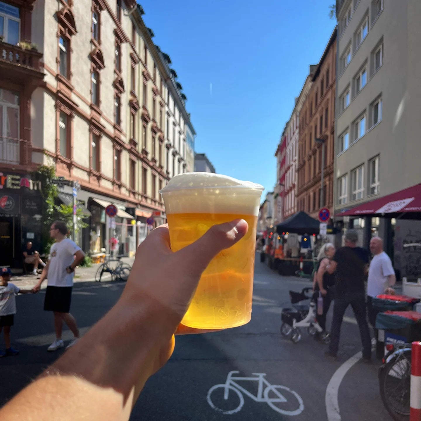 Eine Hand hält einen Plastikbecher mit Bier auf einer belebten Stadtstraße mit Menschen und Marktständen unter blauem Himmel.