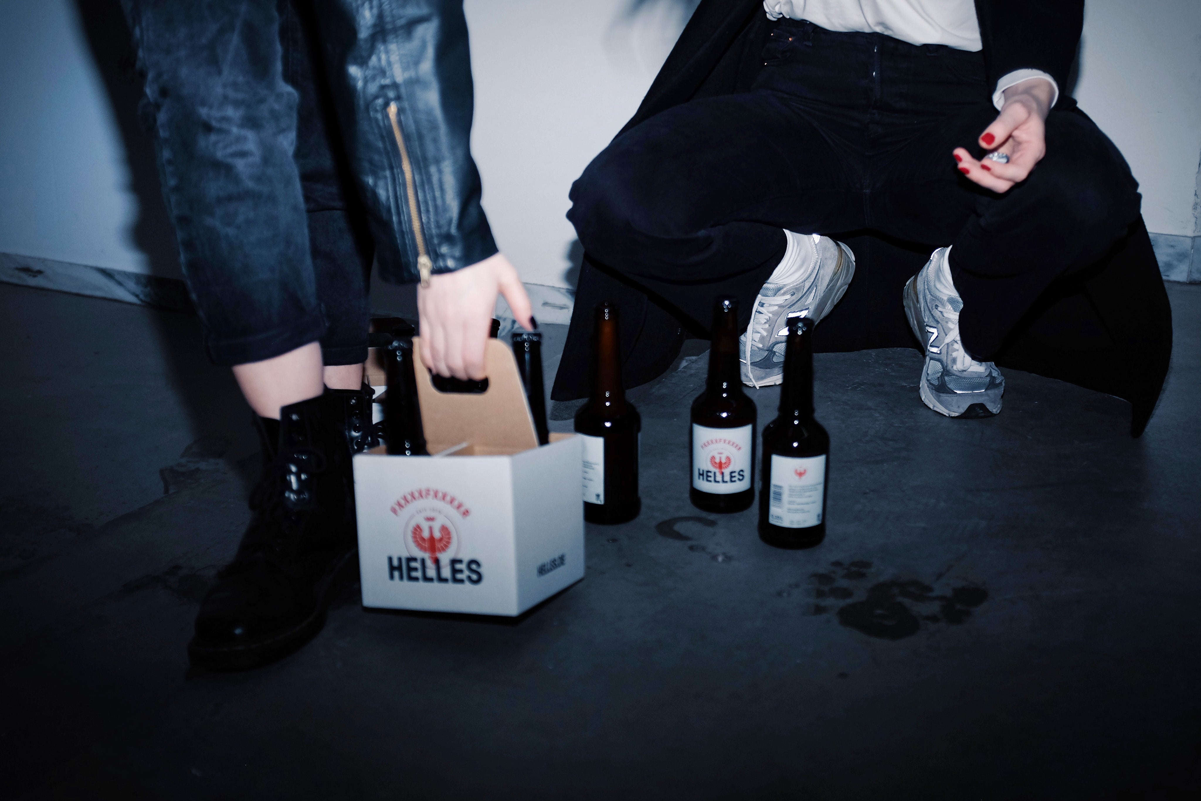 Zwei Personen sitzen mit mehreren Bierflaschen und einem Sechserpack Helles-Bier auf dem Boden, einer greift nach einer Flasche.
