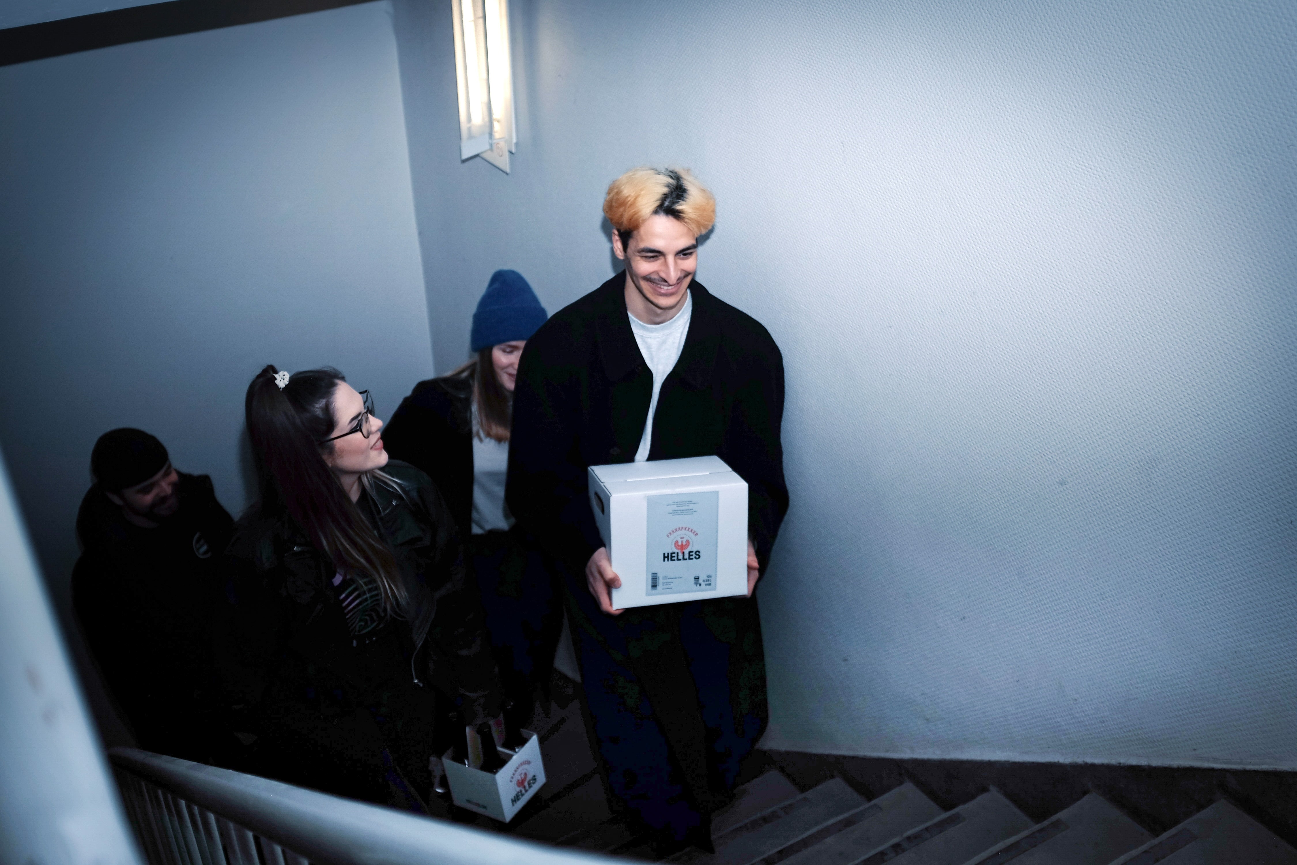 Vier junge Erwachsene gehen eine Treppe hinauf, einer trägt eine Kiste und lächelt, die anderen folgen ihm.