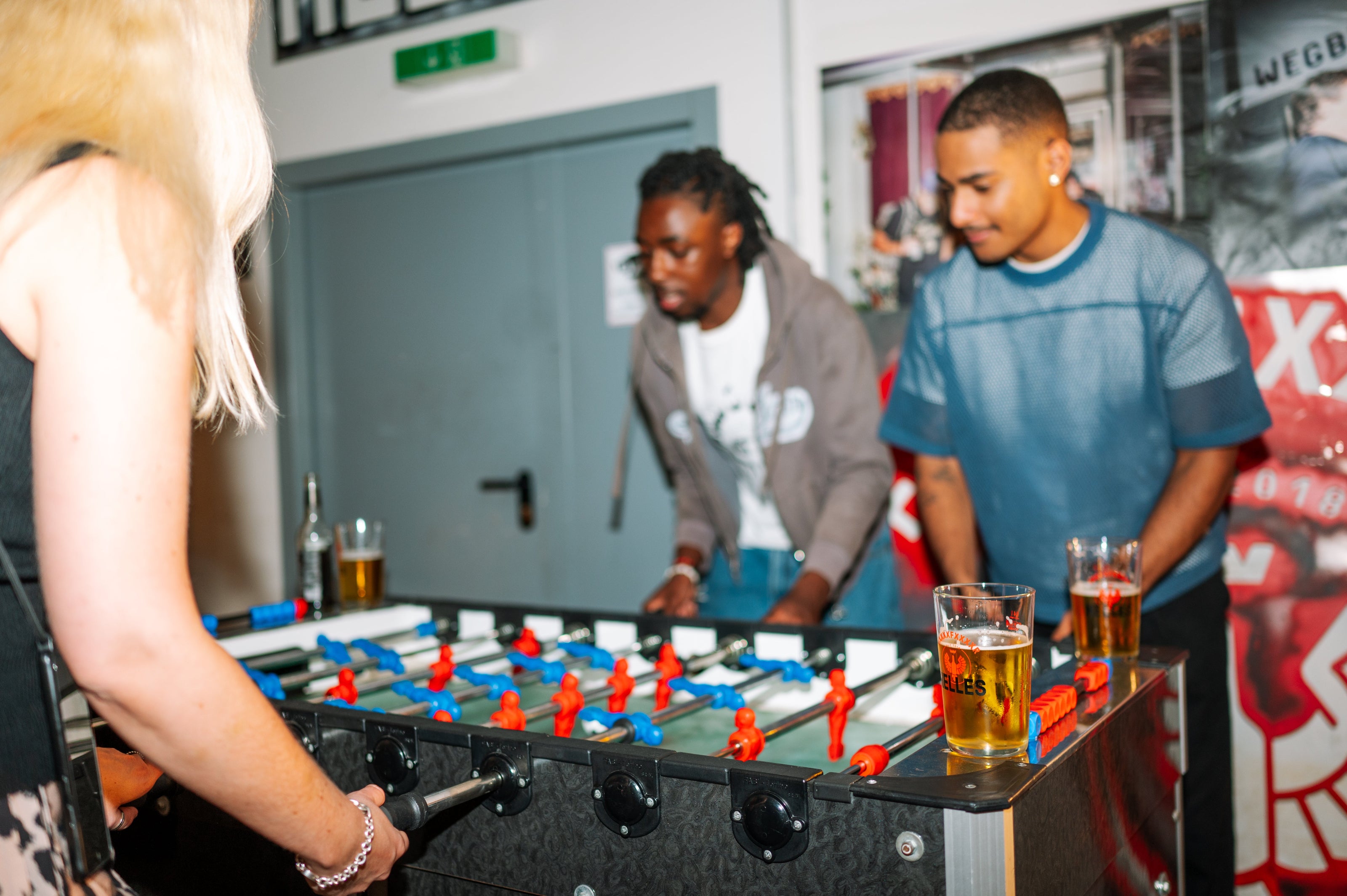 Drei Personen spielen drinnen Tischfußball, mit Getränken auf dem Tisch und Postern an der Wand im Hintergrund.