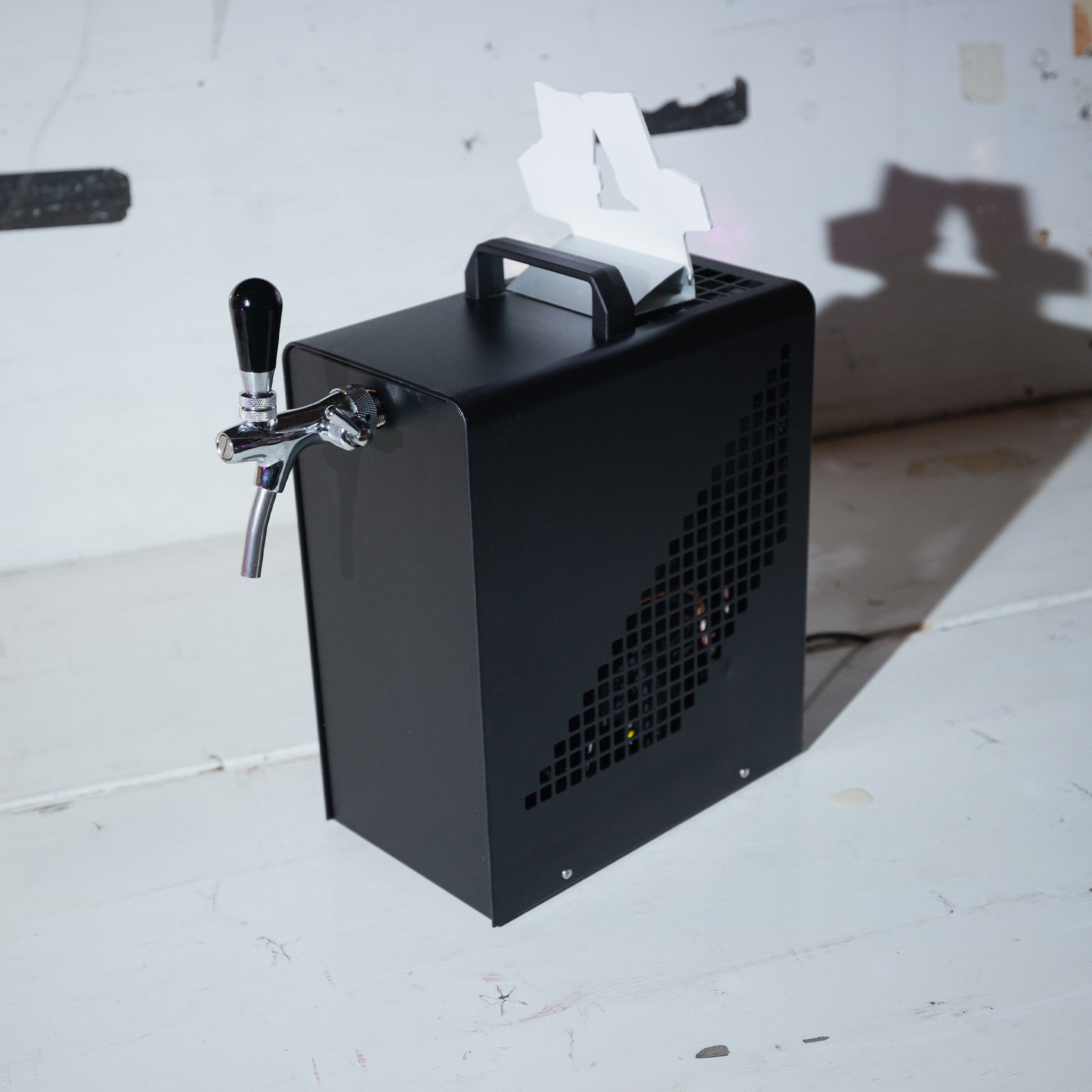 Ein schwarzer Getränkespender mit einem Zapfhahngriff steht auf einer weißen Fläche und wirft einen Schatten an die Wand.