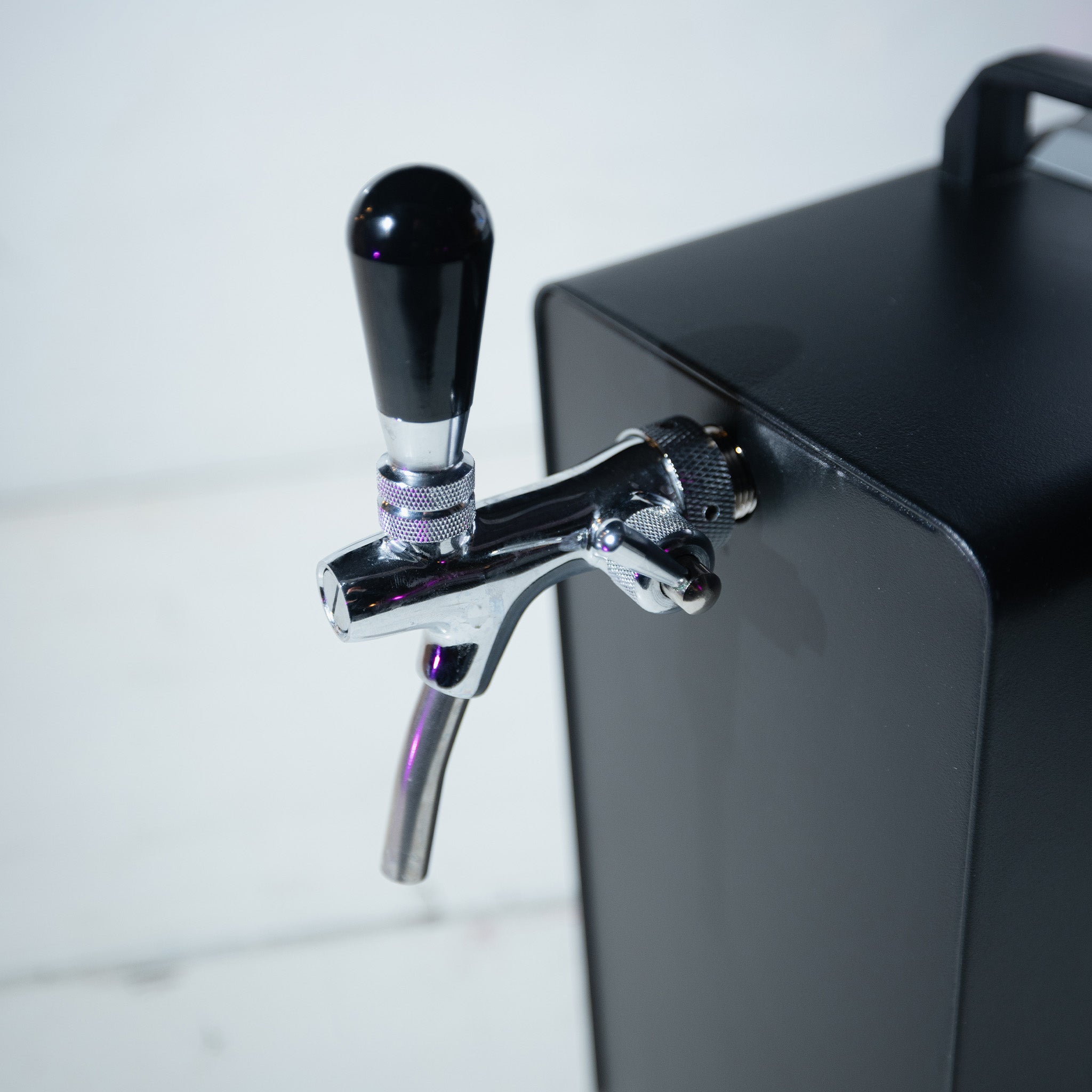 Nahaufnahme eines metallisch glänzenden Zapfhahngriffs, der an einer schwarzen Zapfanlage auf einer weißen Fläche befestigt ist.