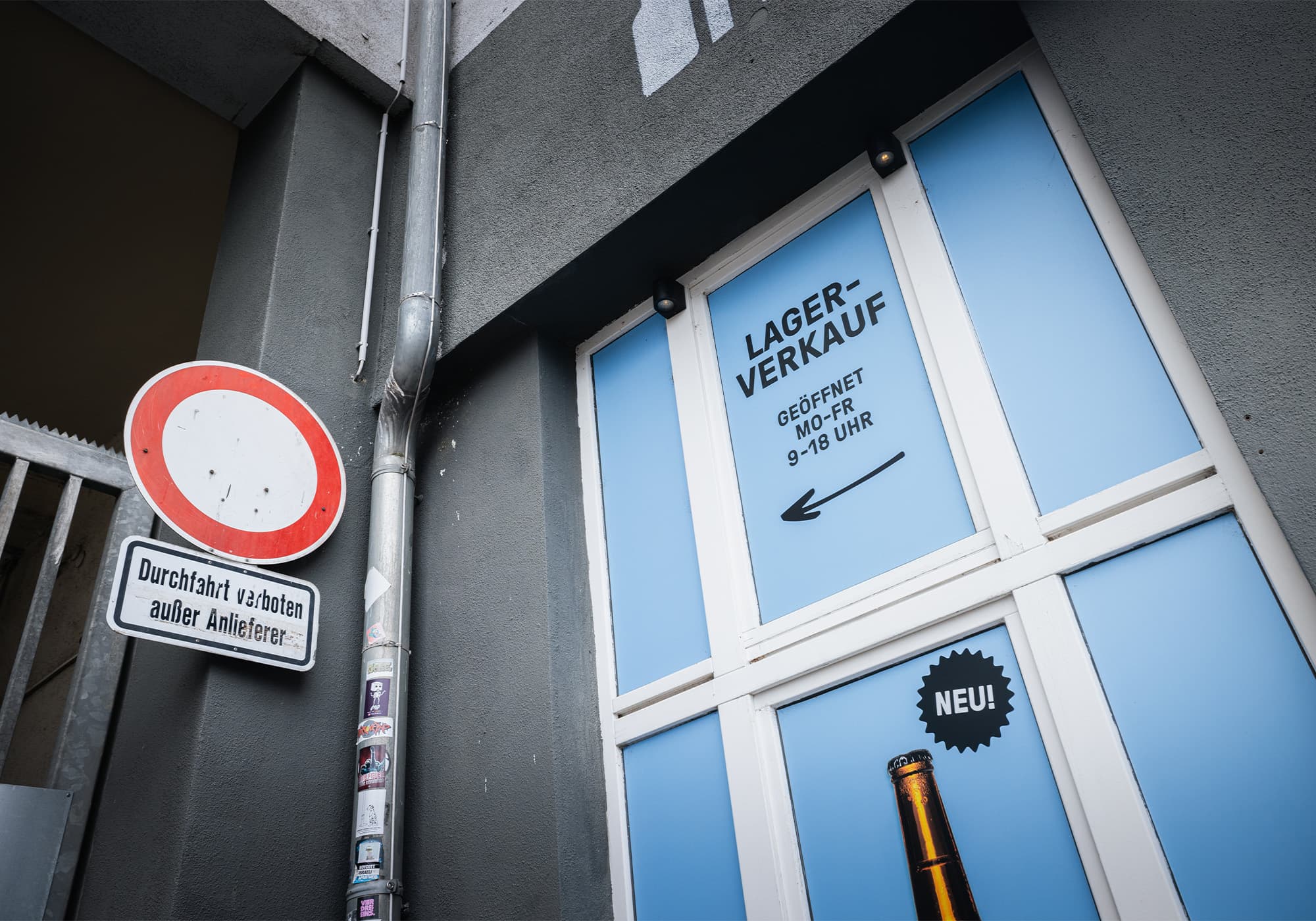 Eine blaue Tür mit dem Schild "Lagerverkauf", ein Straßenschild "Zutritt verboten" und eine teilweise sichtbare Flasche am Boden.