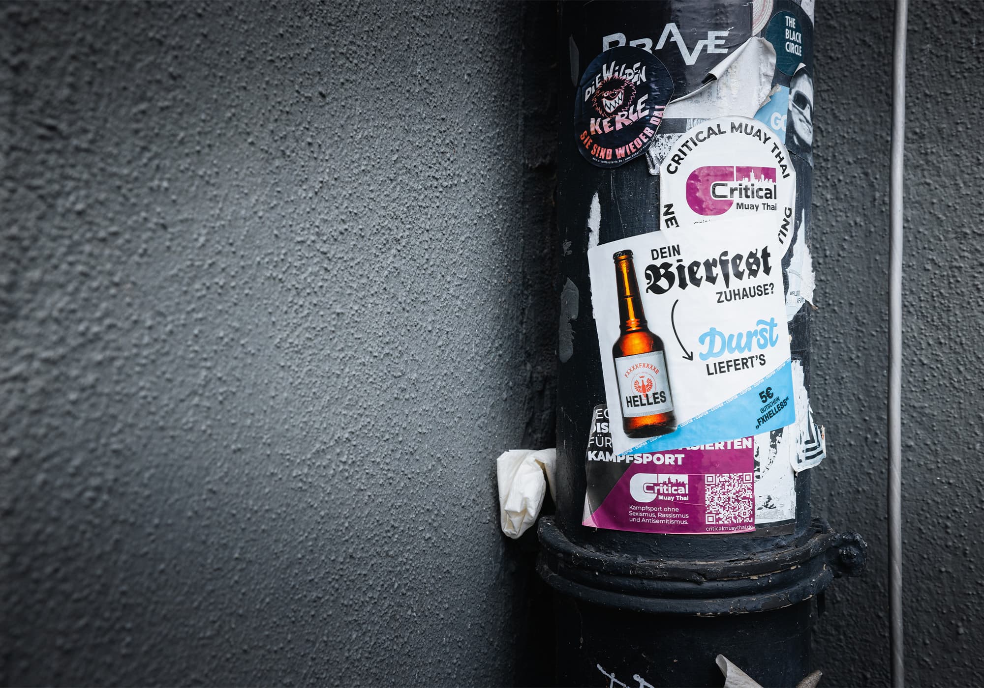 An einer rauen grauen Wand sind Plakate mit deutschem Text und einer Bierflasche an einer schwarzen Stange befestigt.