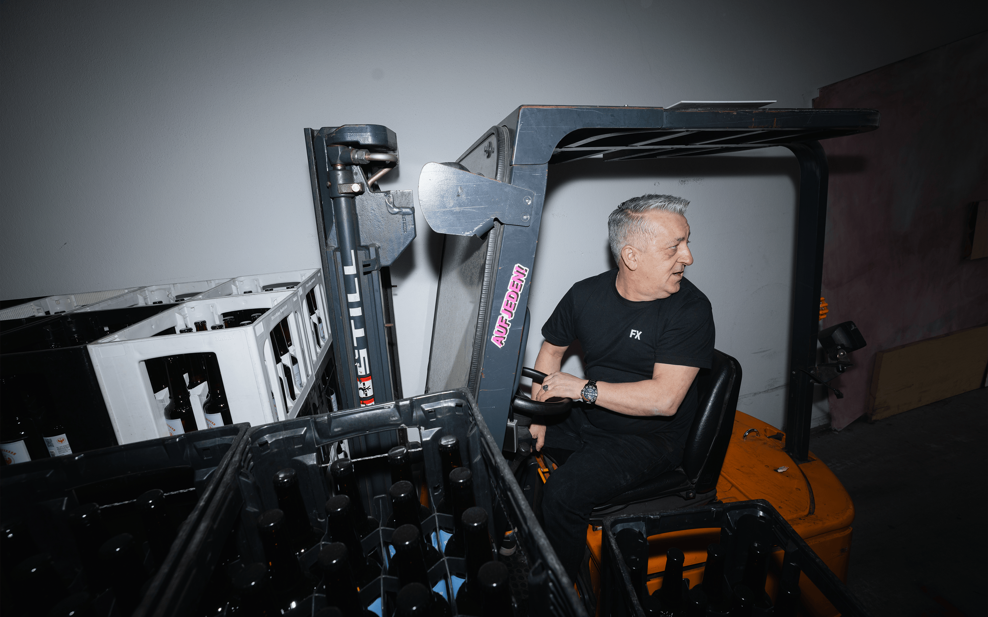 Ein Mann in einem schwarzen Hemd fährt einen Gabelstapler neben Kisten mit leeren Glasflaschen in einer Halle.