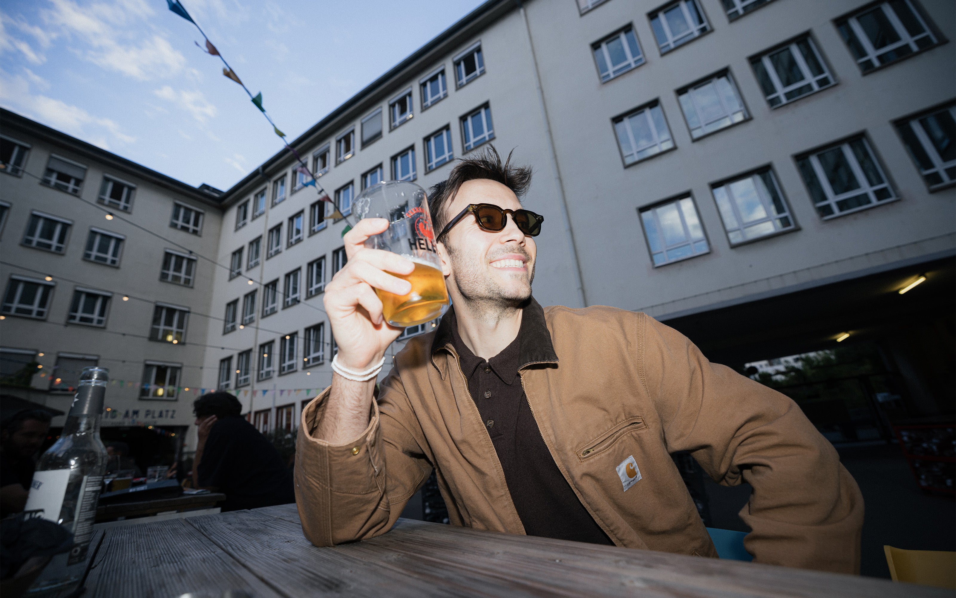 Mann mit Sonnenbrille und brauner Jacke lächelt, während er an einem Tisch im Freien in einem städtischen Innenhof ein Getränk zu sich nimmt.
