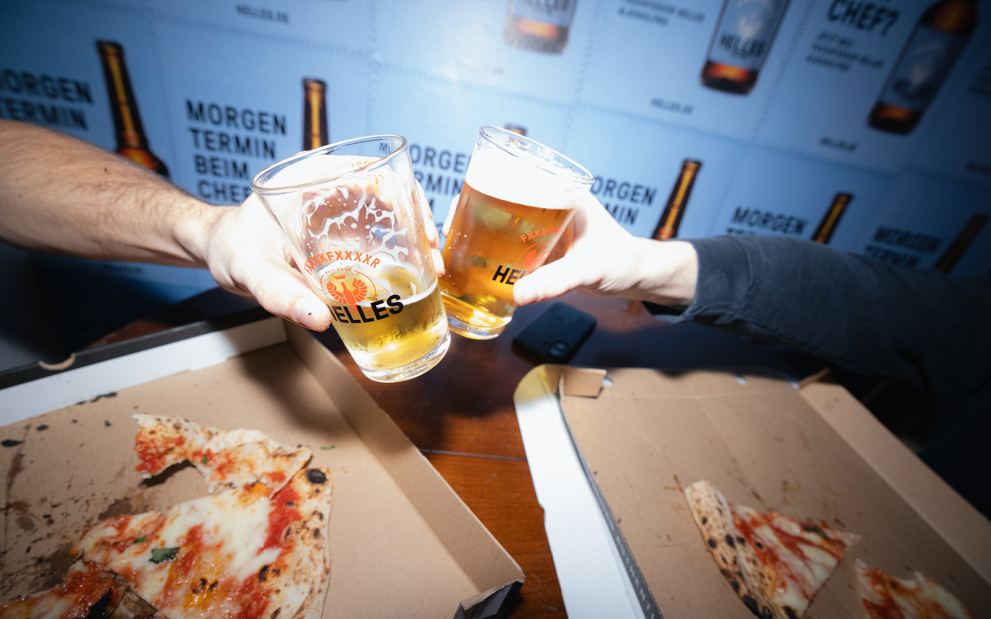 Zwei Personen stoßen mit Biergläsern über einem Tisch mit Pizza in einer Schachtel an, vor einem Hintergrund mit Bierlogos.