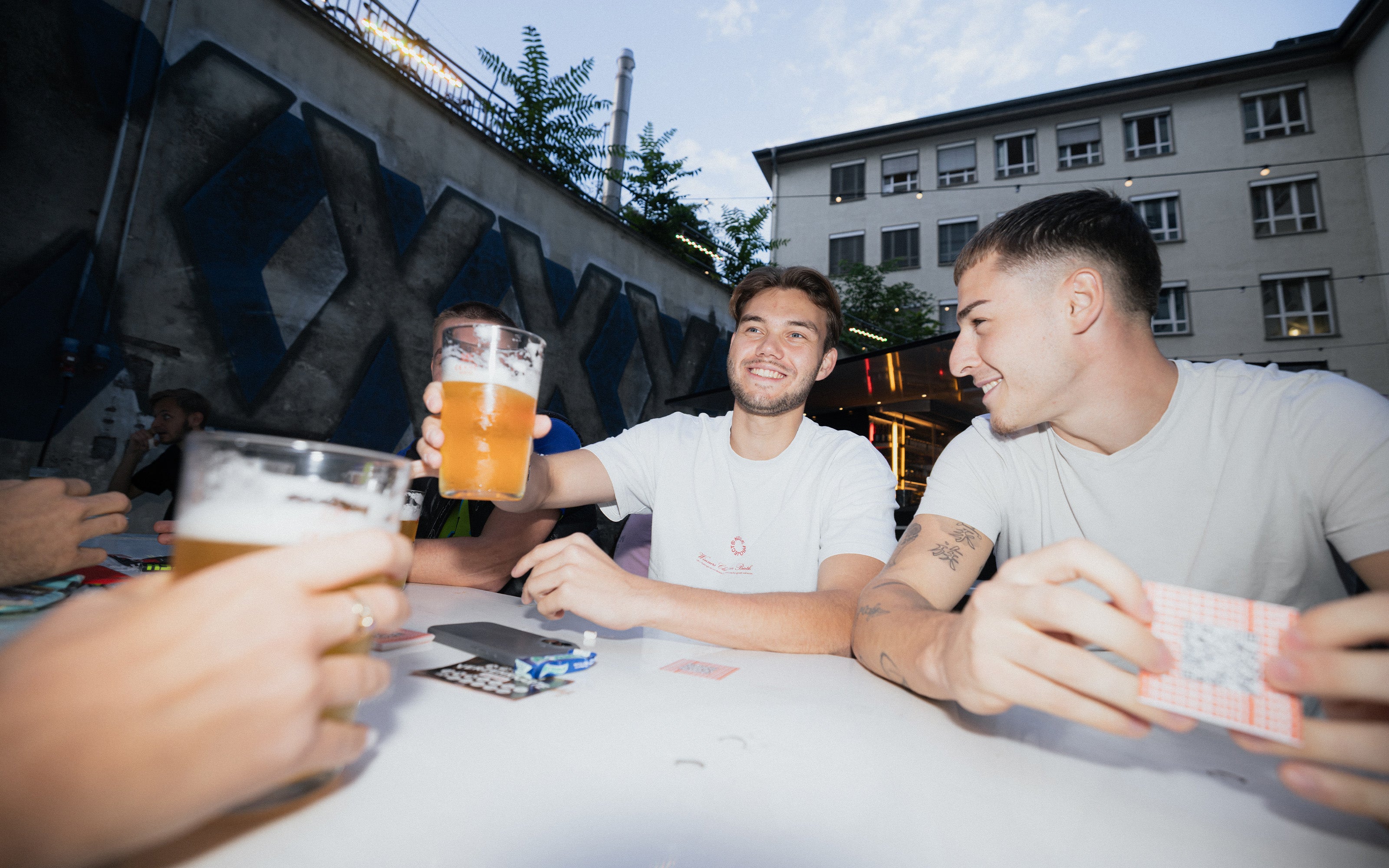 Zwei junge Männer sitzen im Freien an einem Tisch, lächeln, halten Biergläser in der Hand und spielen Karten.