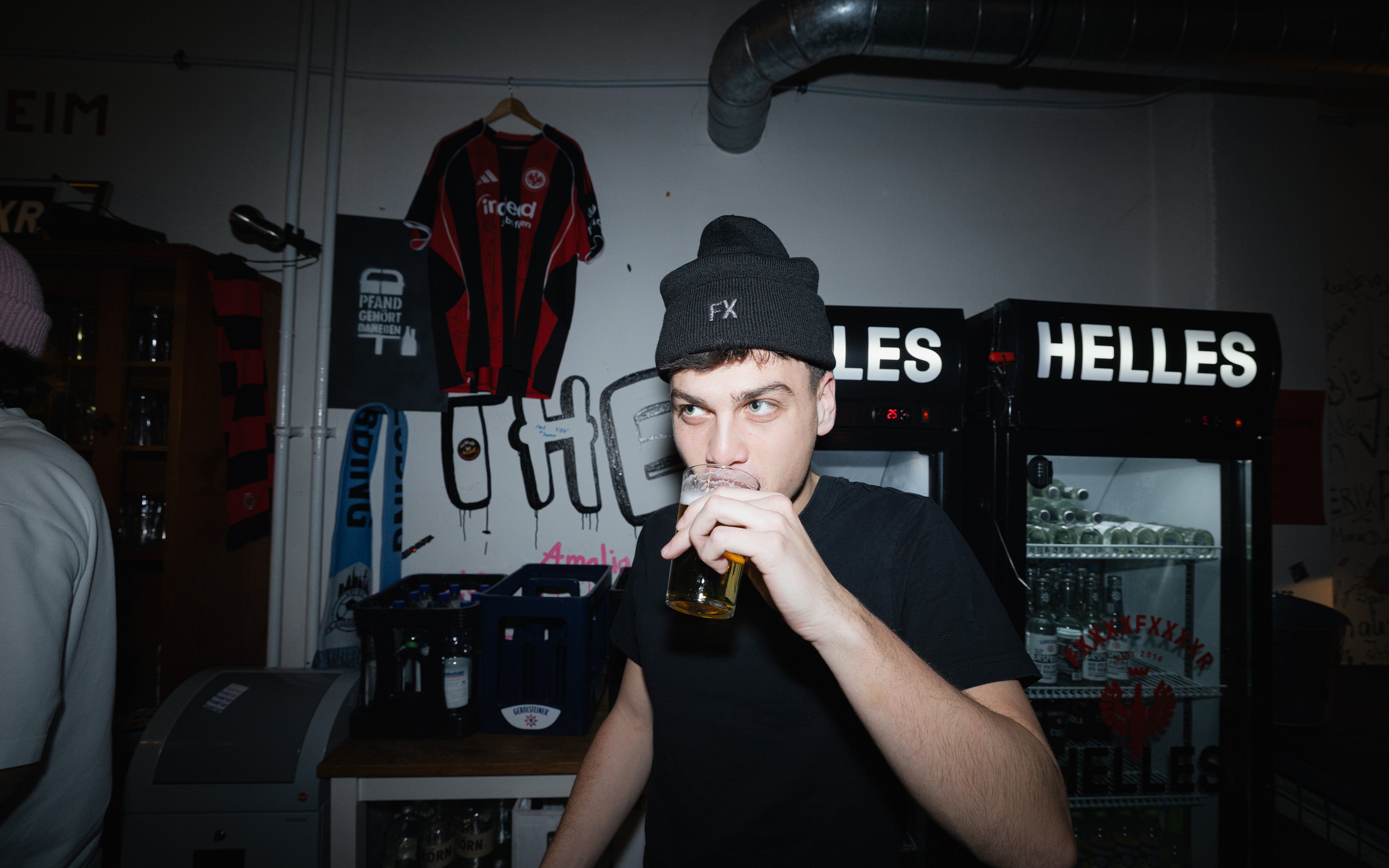 Ein Mann mit schwarzer Mütze trinkt aus einem Glas in einer Bar mit Trikots und Kühlschränken mit der Aufschrift HELLES im Hintergrund.