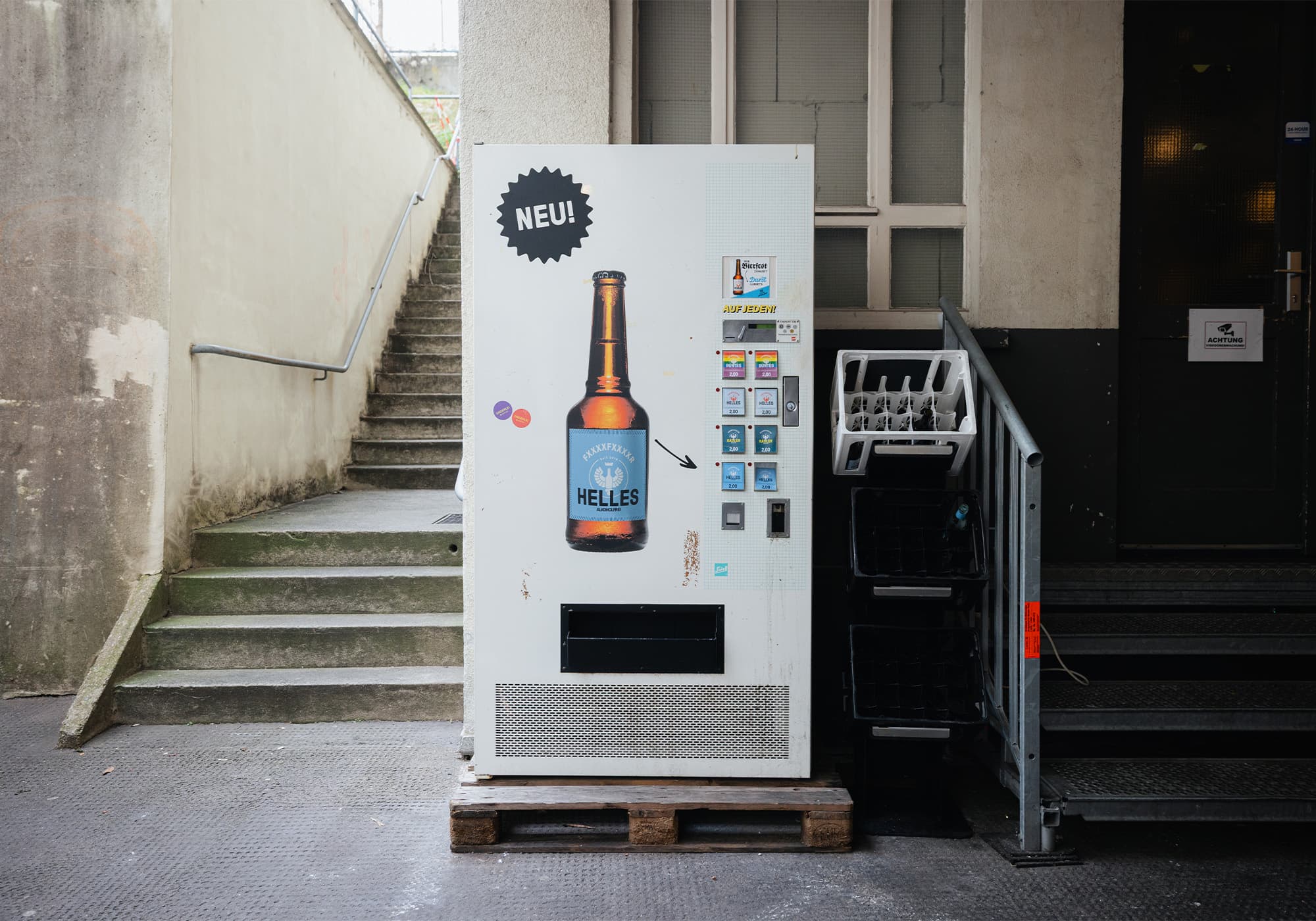 Ein weißer Verkaufsautomat mit einer großen Bierflaschengrafik steht neben einer Treppe in einem städtischen Außenbereich.