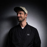 Mann in FX Jacke mit reflektierendem Print und beiger Cap vor dunklem, gradienten Hintergrund.
