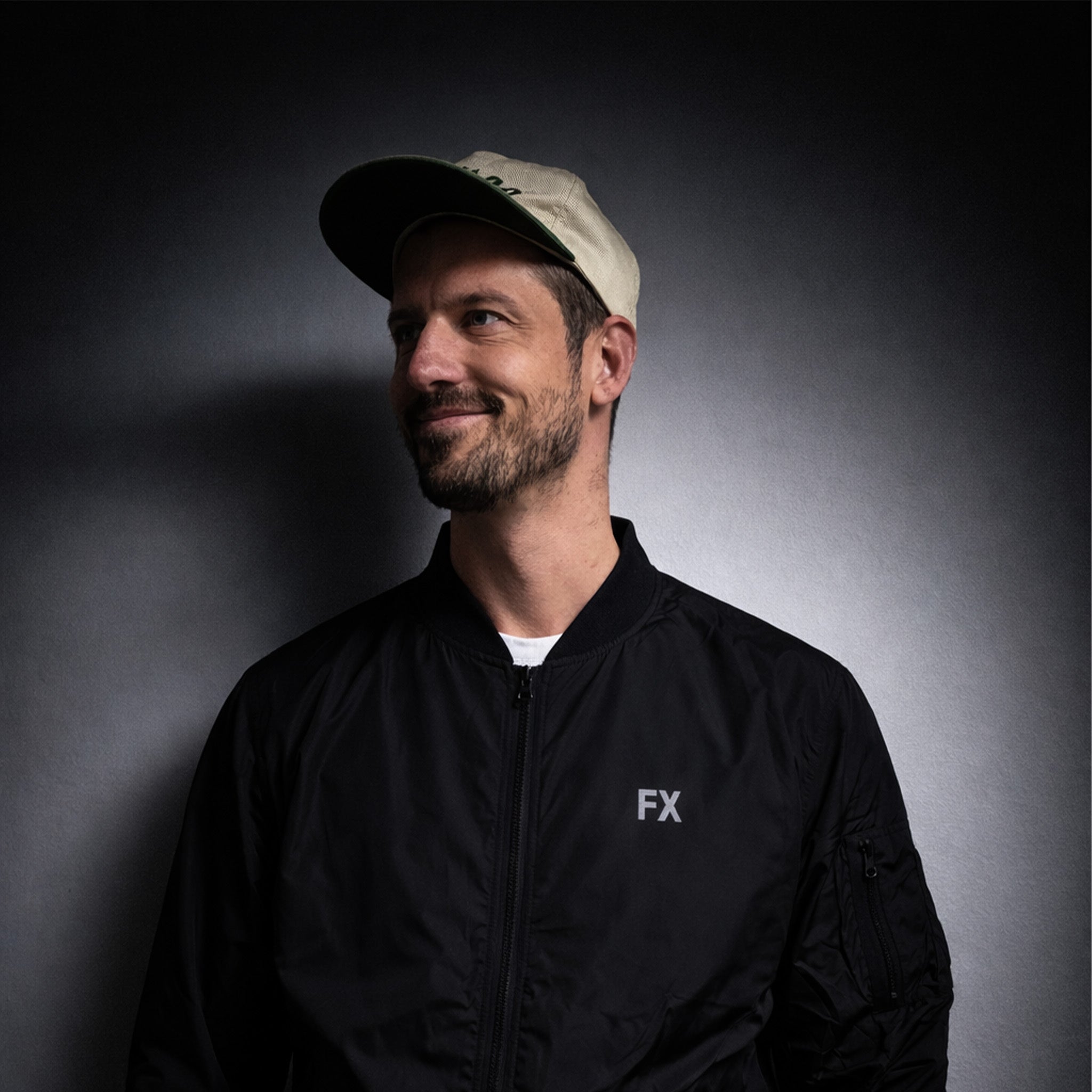 Mann in FX Jacke mit reflektierendem Print und beiger Cap vor dunklem, gradienten Hintergrund.