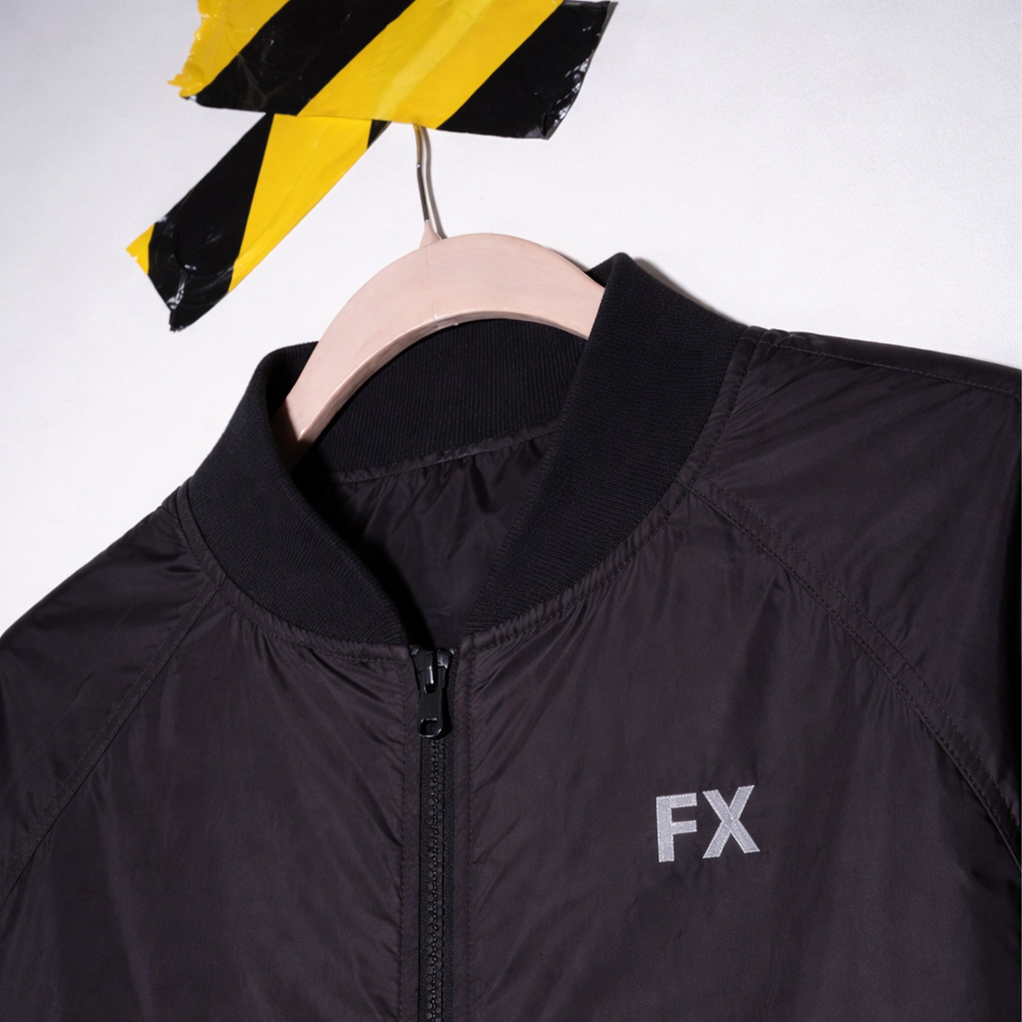FX Jacke mit Stickerei auf der Brust hängt am Holzkleiderbügel vor weißer Wand mit Caution Tape darüber.