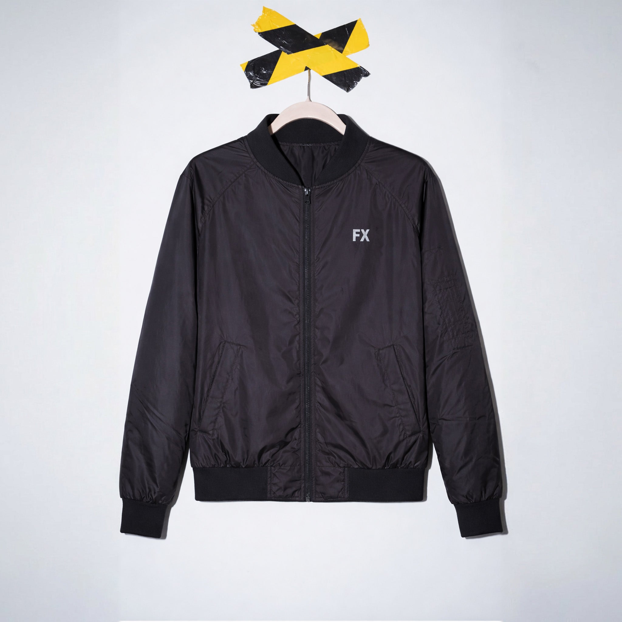 FX Jacke mit reflektierendem Print hängt an einem Holzkleiderbügel unter schwarz-gelbem Band an der Wand.