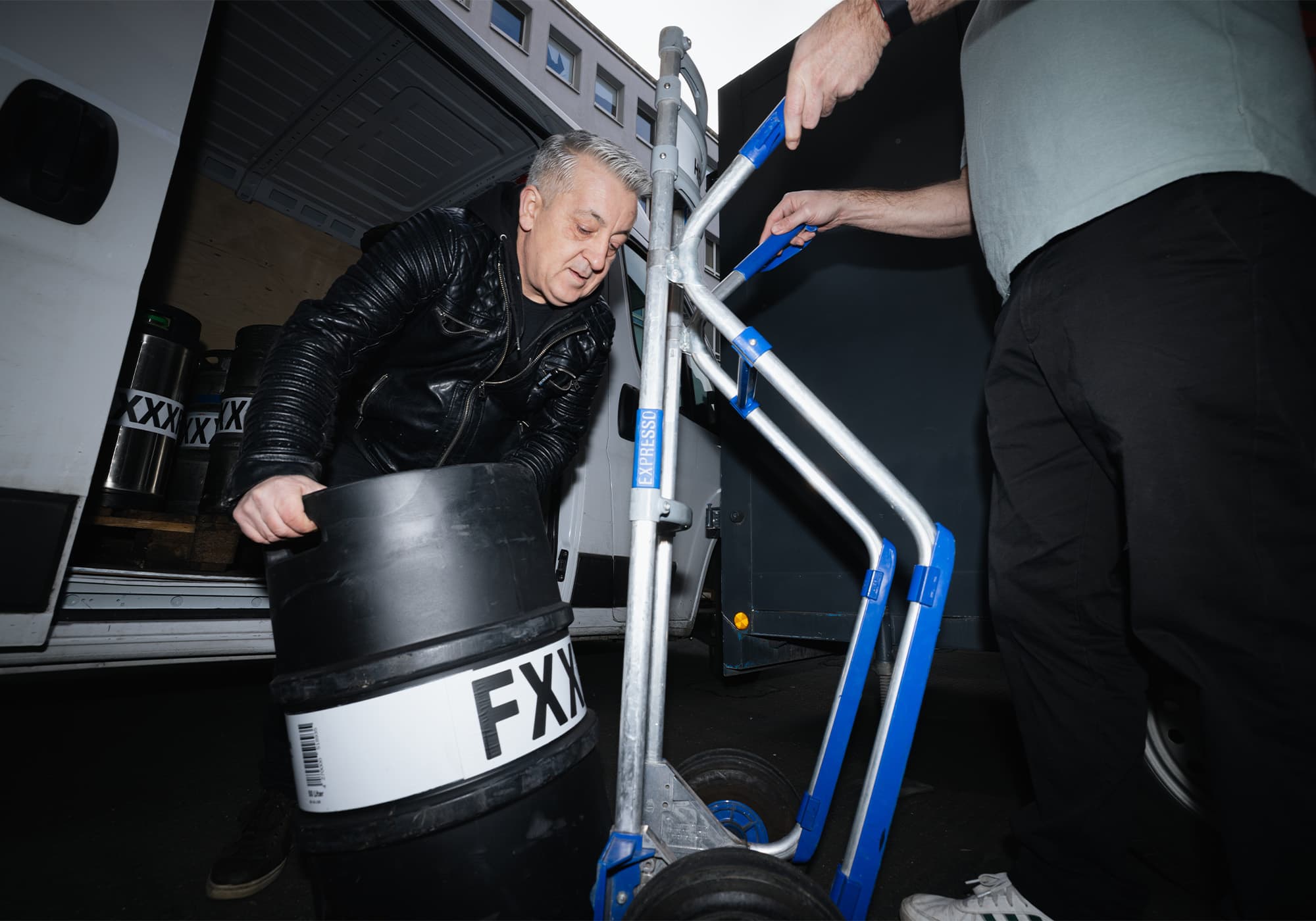 Zwei Männer laden große Fässer mit FX-Etikett auf einen blauen Handwagen neben einem Lieferwagen.