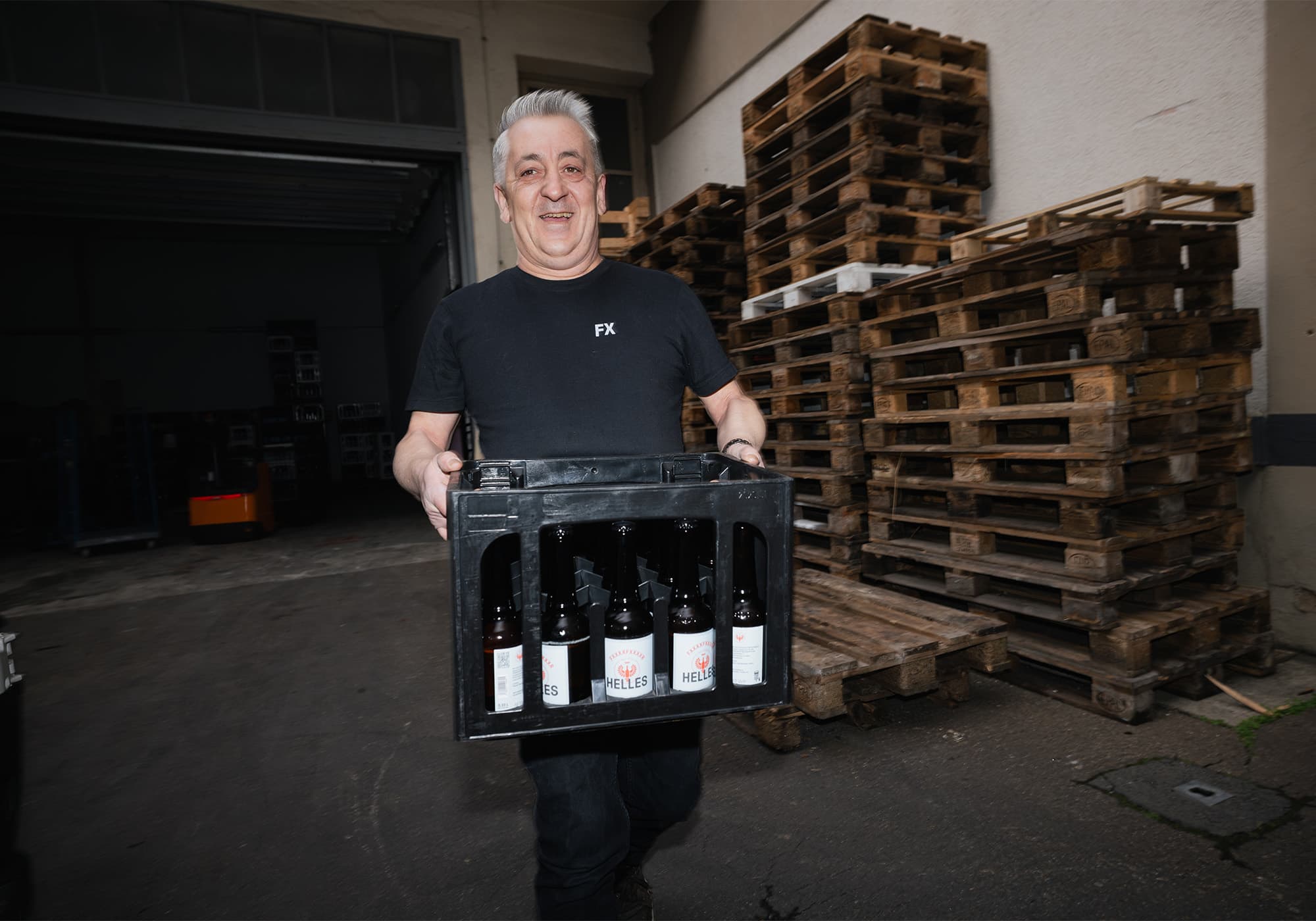 Ein lächelnder Mann in einem schwarzen Hemd trägt eine Kiste mit Getränkeflaschen neben gestapelten Holzpaletten ins Freie.