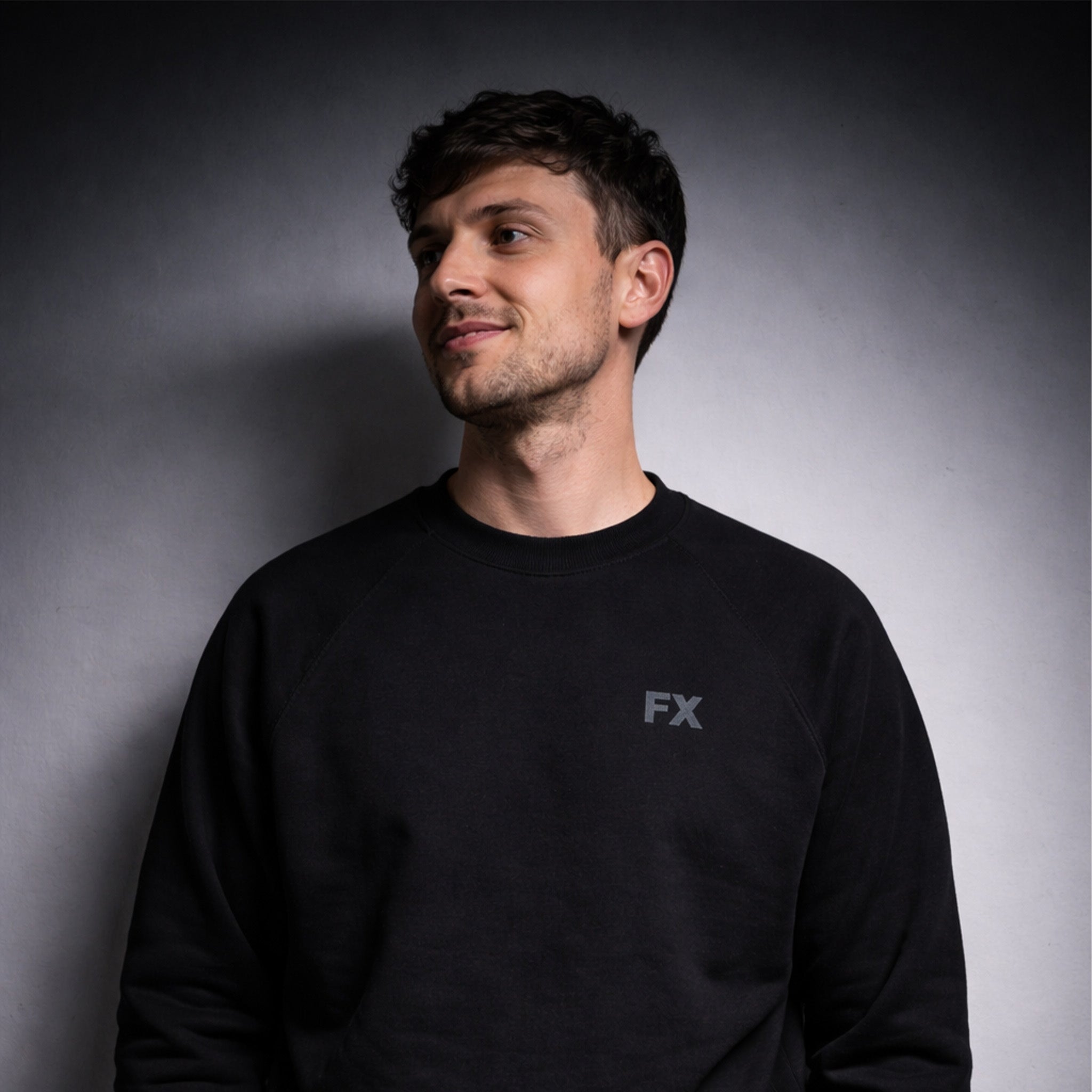 Ein Mann trägt den FX Pulli in Schwarz mit reflektierendem Print und lächelt vor grauem Hintergrund.