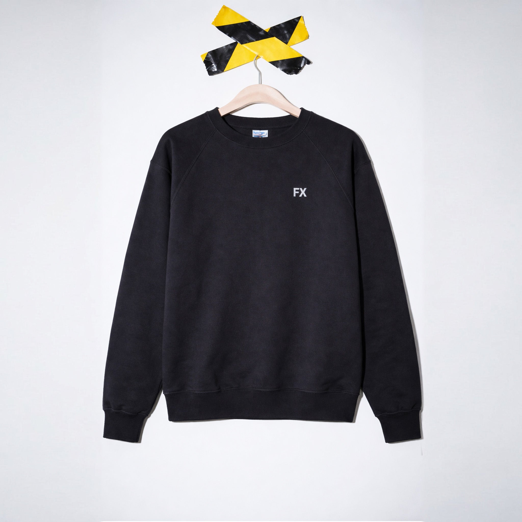 FX Pulli: Schwarzes Crewneck-Sweatshirt mit Raglanärmeln, reflektierender FX-Druck, auf Bügel unter gelb-schwarzem Band.