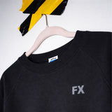 Der FX Pulli mit Raglanärmeln und FX Schriftzug hängt an einem Holzbügel unter schwarz-gelb gestreiftem Band.