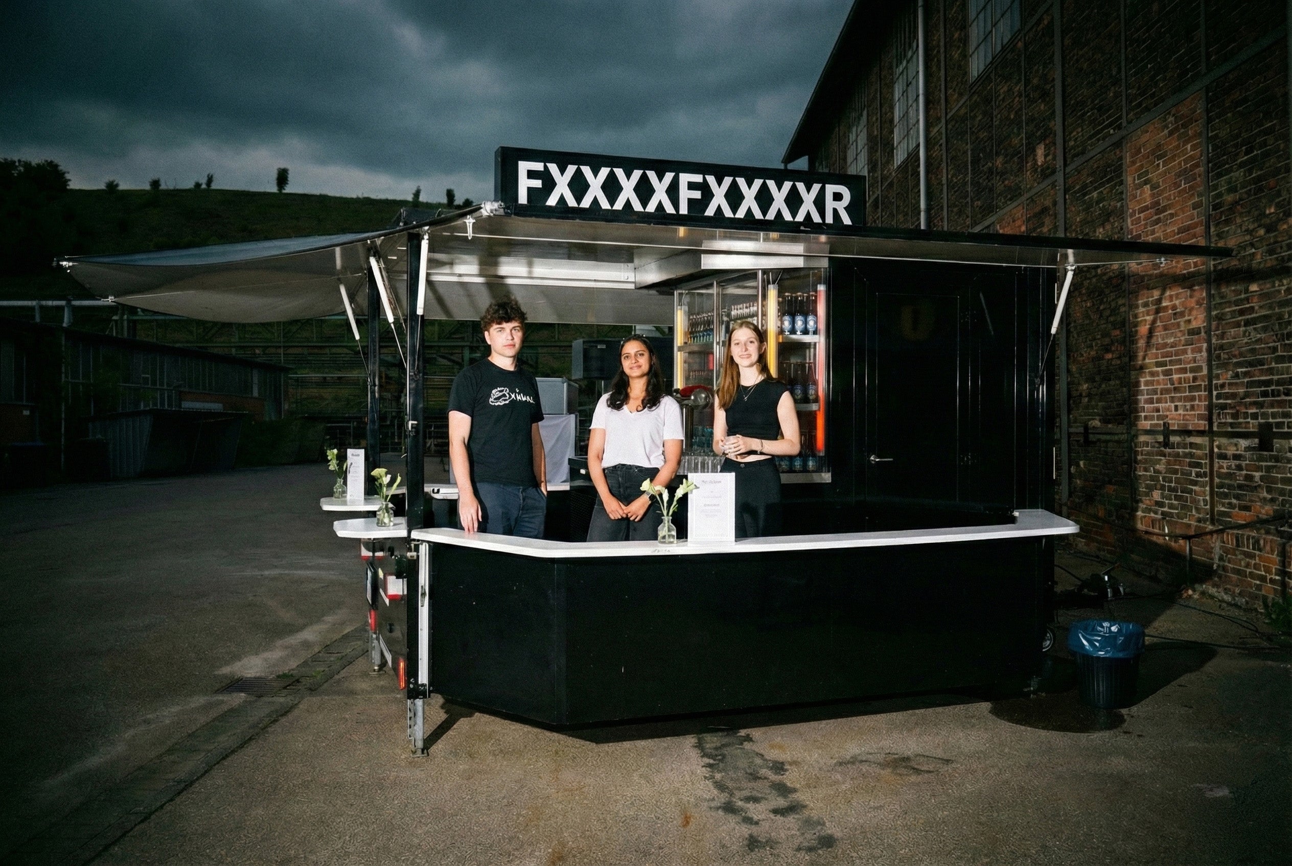 Drei Personen stehen an einem bewölkten Tag hinter einem schwarzen Imbissstand mit einem Schild mit der Aufschrift FXXXXFXXXXR.