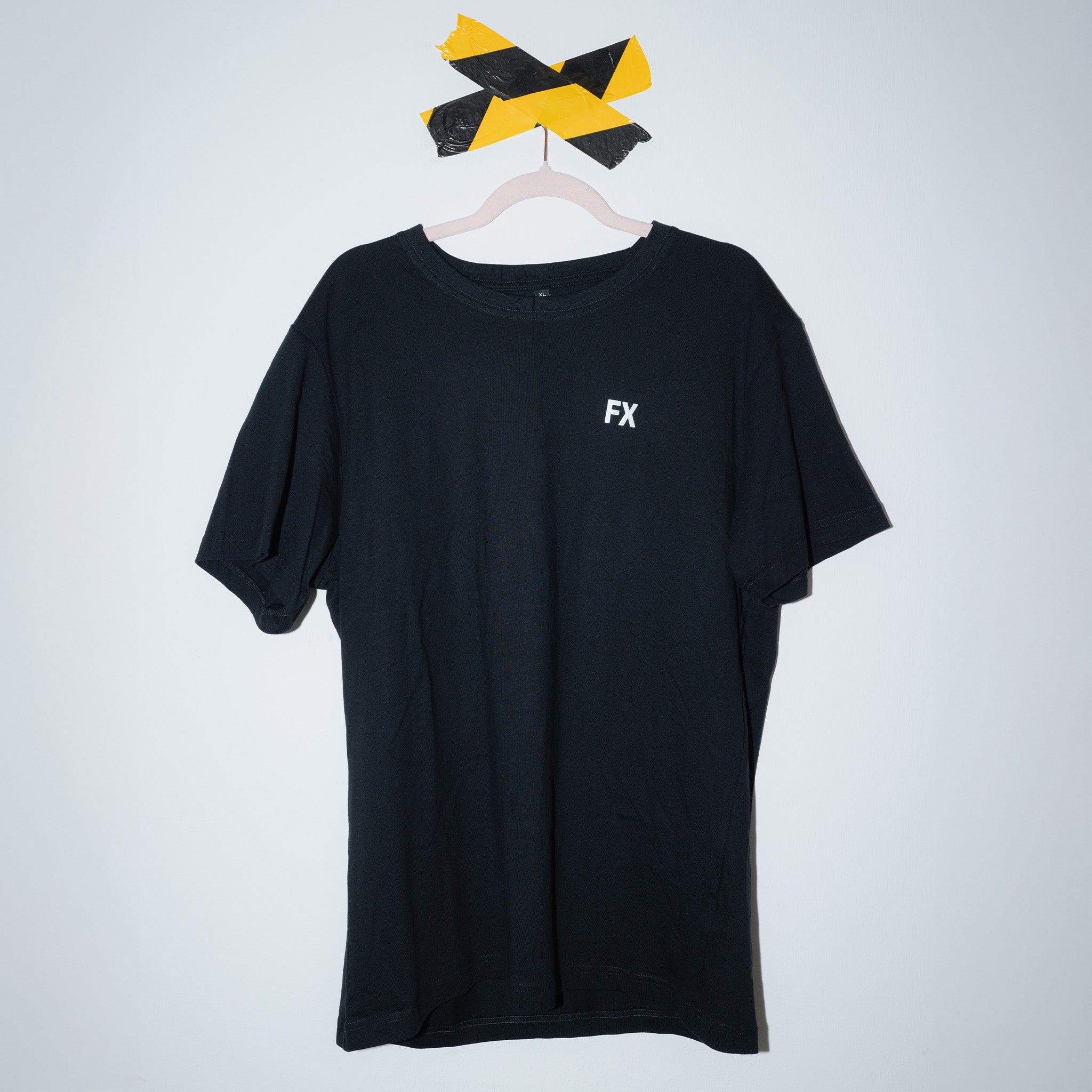 Das FX T-Shirt von Frankfurter Brauunion aus Oeko-Tex Baumwolle hängt an einem Kleiderbügel, fixiert mit schwarz-gelbem Tape vor weißer Wand. Auf der linken Brust befindet sich ein reflektierender "FX"-Print.