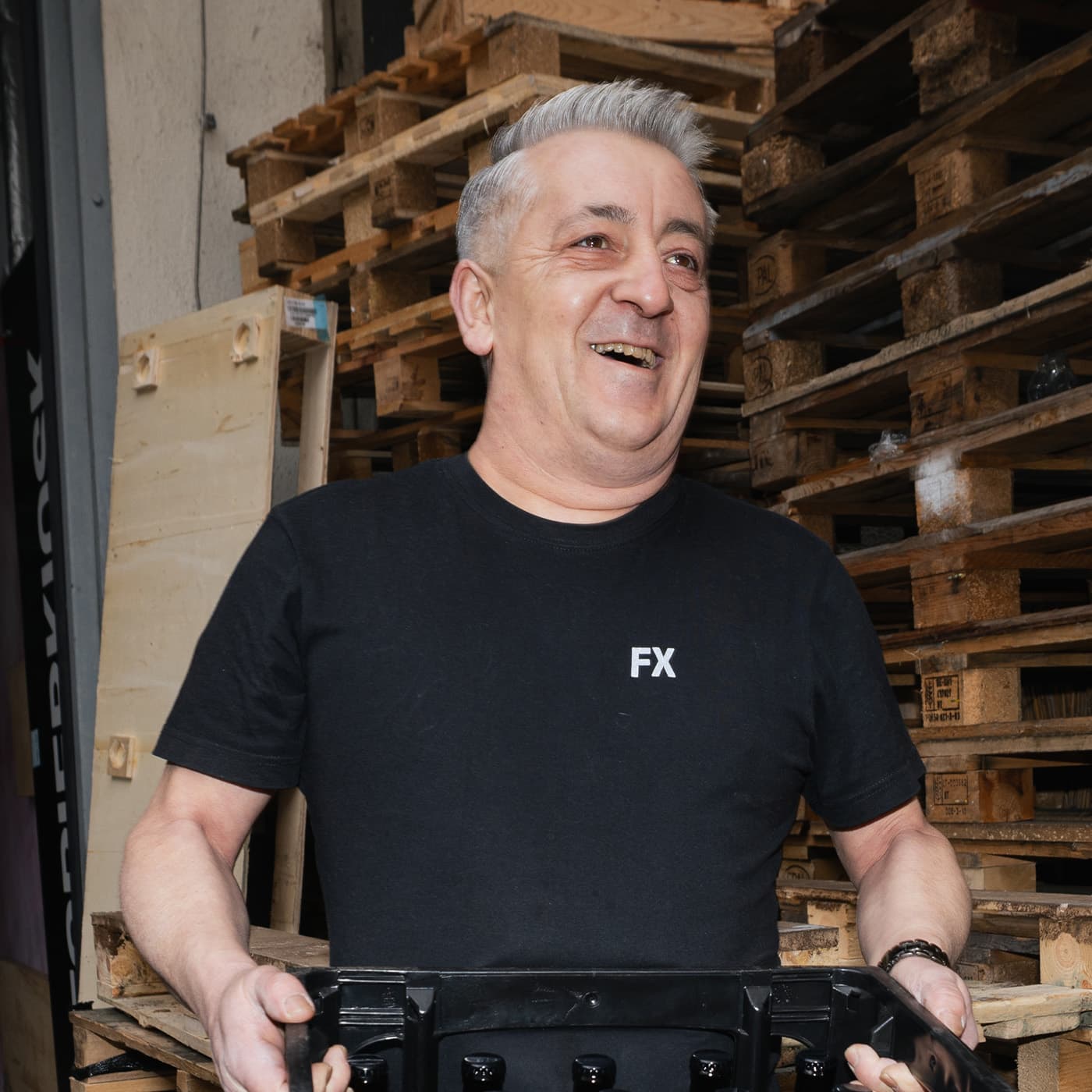 Ein lächelnder Mann mit kurzen grauen Haaren hält einen schwarzen Container und steht vor gestapelten Holzpaletten.
