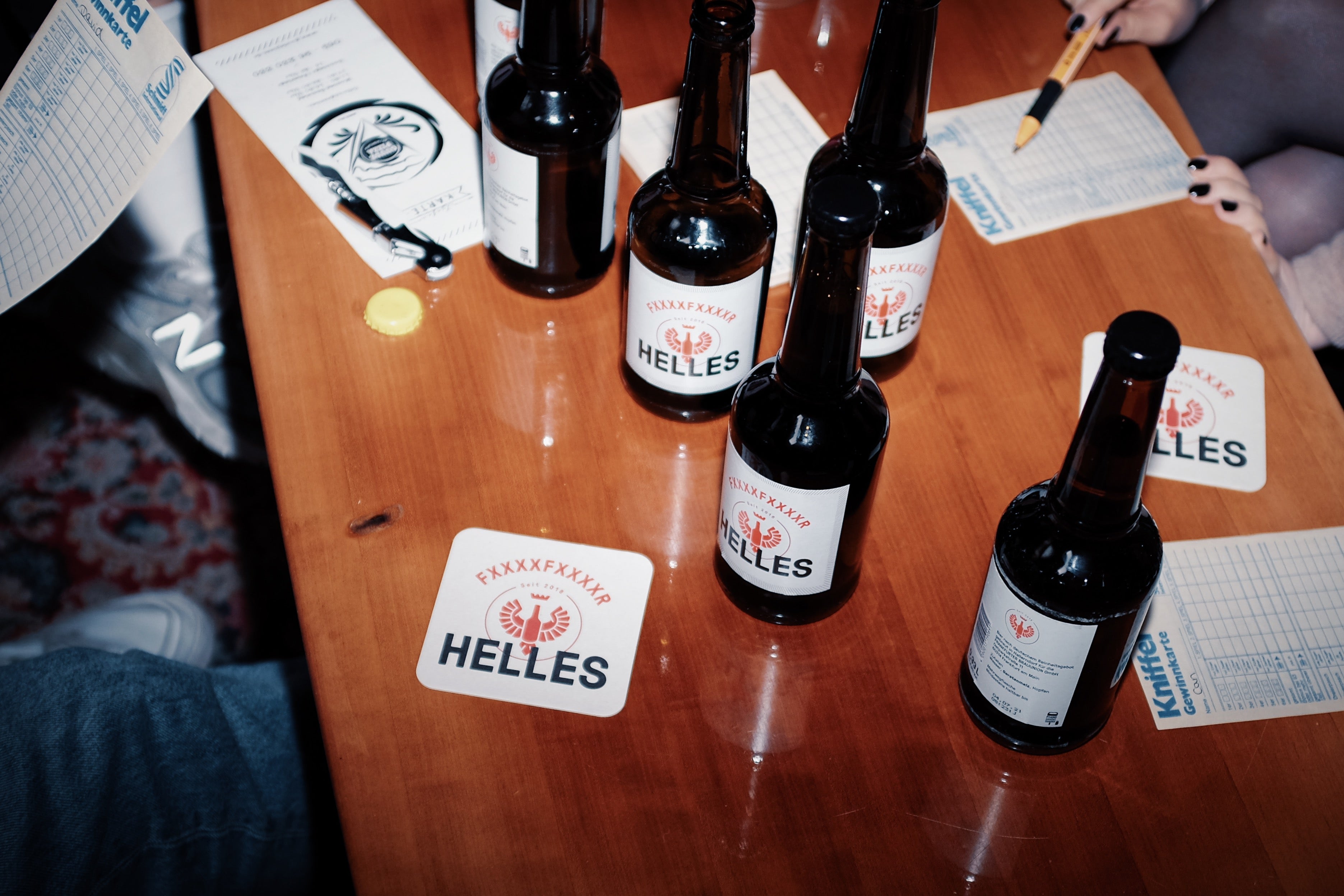 Mehrere Flaschen Helles-Bier auf einem Holztisch mit Untersetzern, daneben Leute, die Formulare ausfüllen.