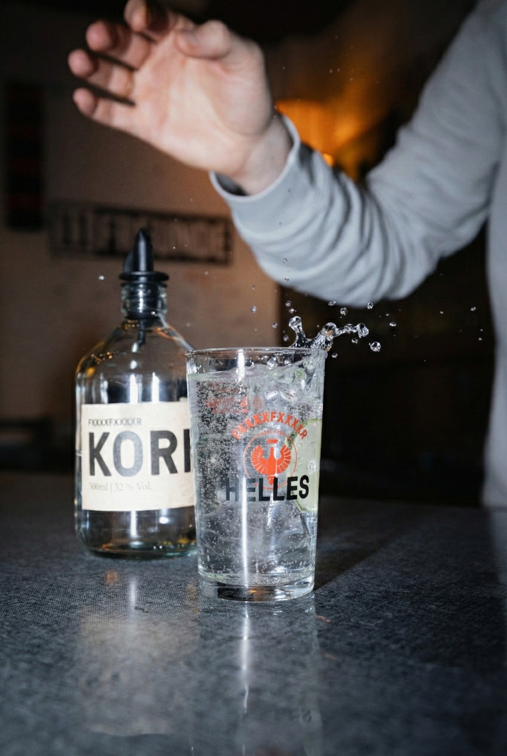 Eine Hand spritzt Wasser in ein Glas neben einer Flasche mit der Aufschrift KORI auf einer reflektierenden Oberfläche.