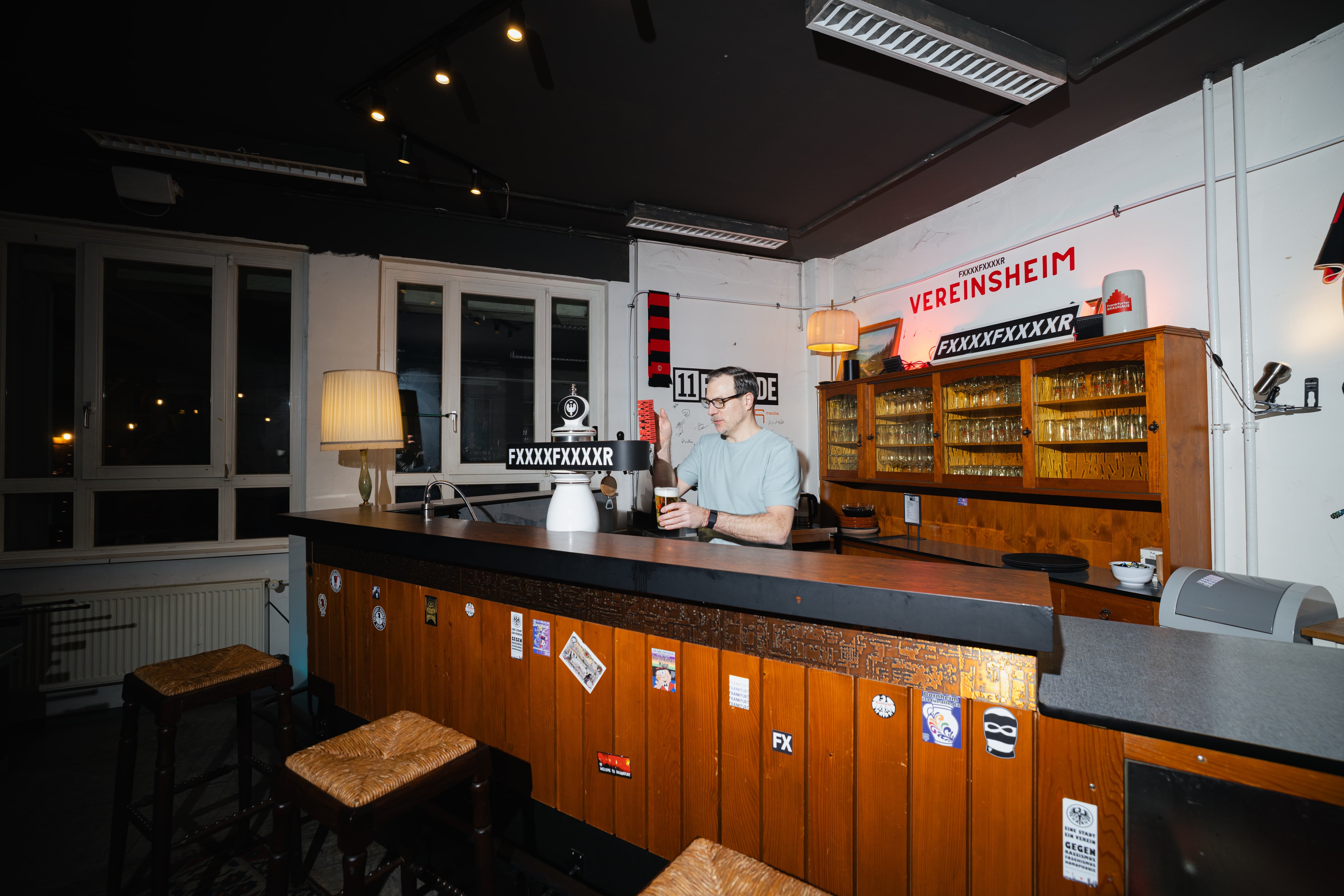 Ein Mann sitzt allein an einem Bartresen in einem schwach beleuchteten Raum mit Holzhockern und Glasregalen hinter ihm.