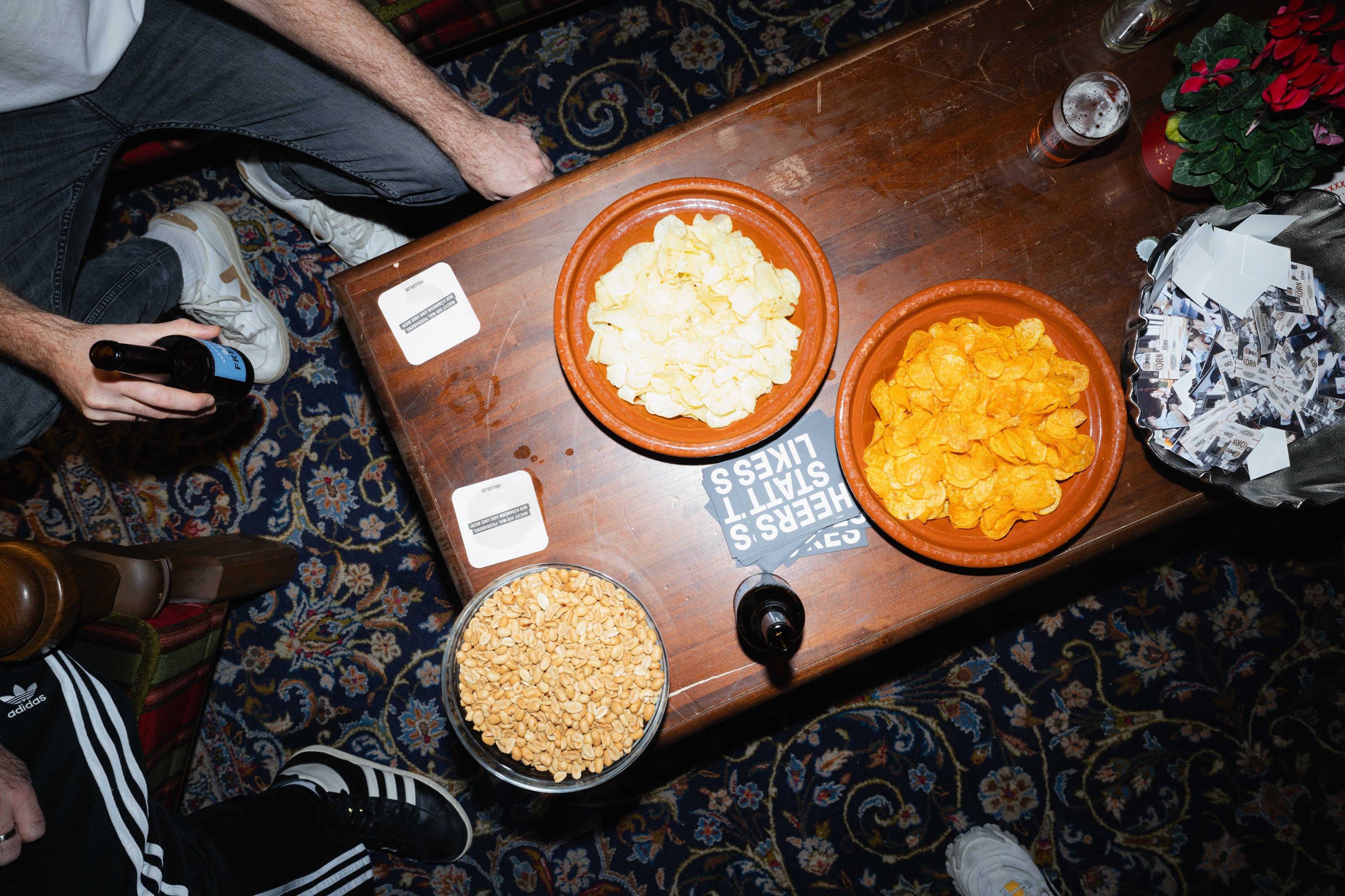 Ein Tisch mit Chips, Erdnüssen, Getränken und Untersetzern, umgeben von Menschen, von oben gesehen.