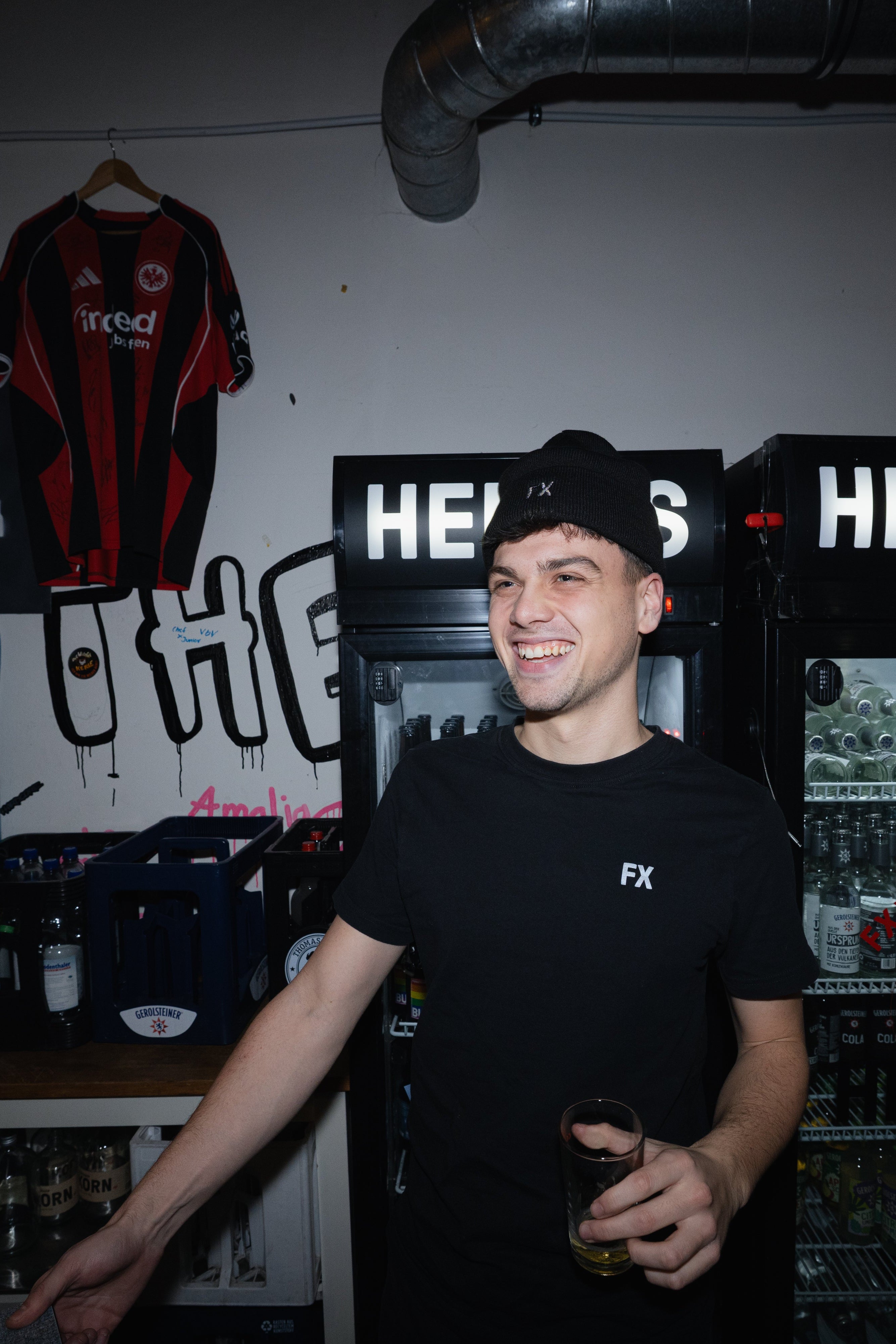 Ein lächelnder Mann in schwarzem Hemd und Mütze steht mit einem Getränk in der Hand vor den Kühlschränken einer Bar.