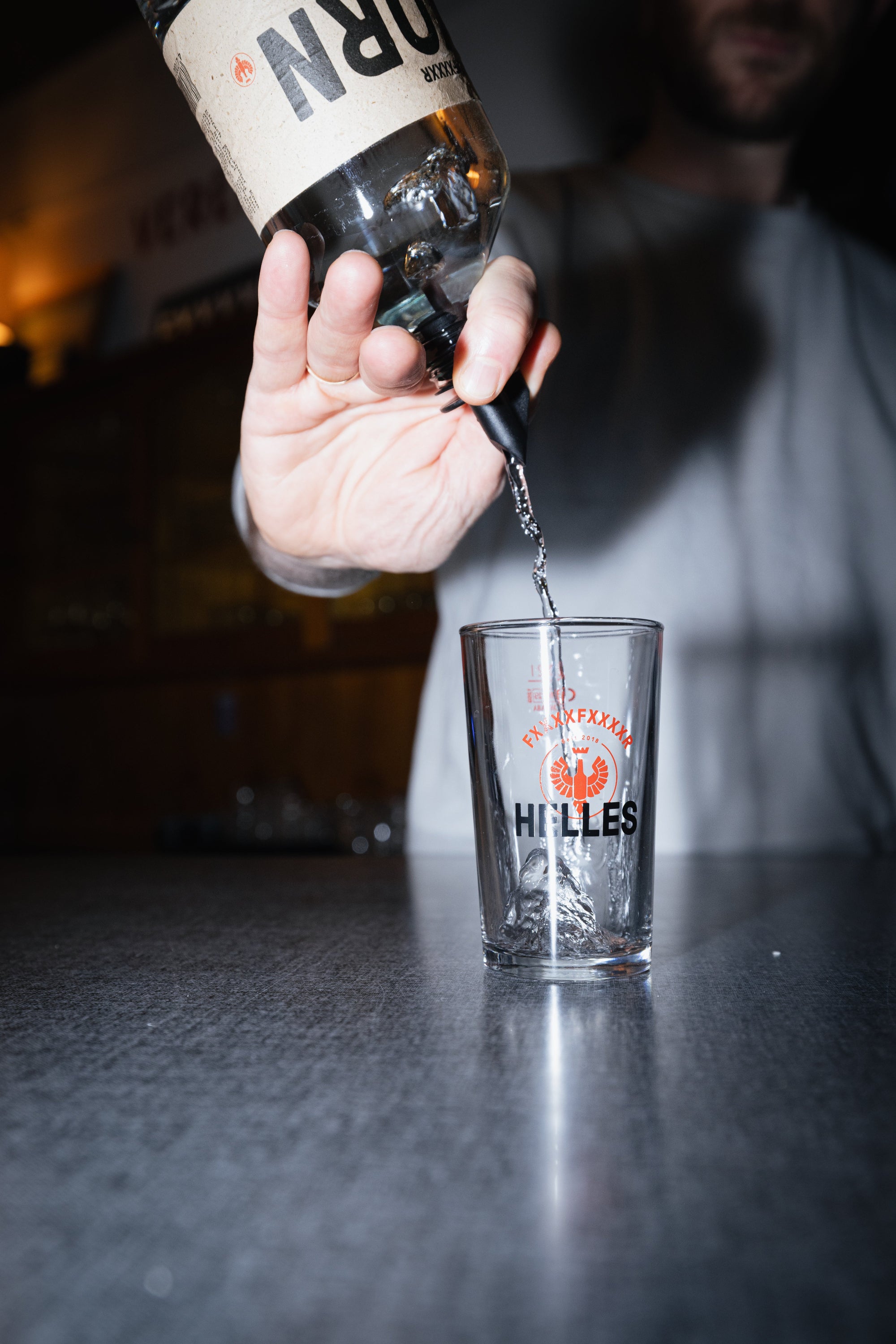 Eine Person gießt klaren Schnaps aus einer Flasche in ein Glas mit der Aufschrift HILLES auf einem Tresen.