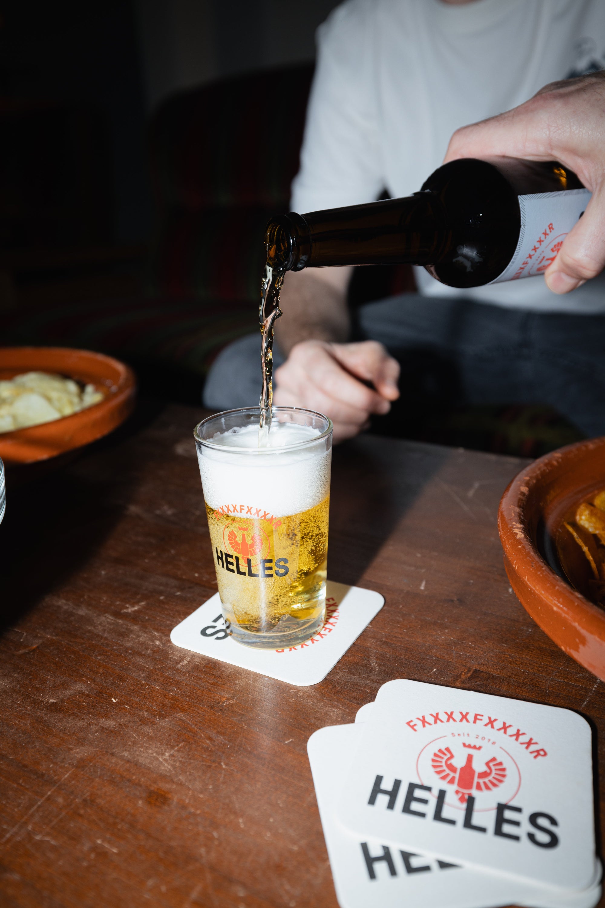 Eine Hand gießt Helles-Bier aus einer Flasche in ein Glas auf einem Untersetzer, daneben stehen Snacks in Schalen.