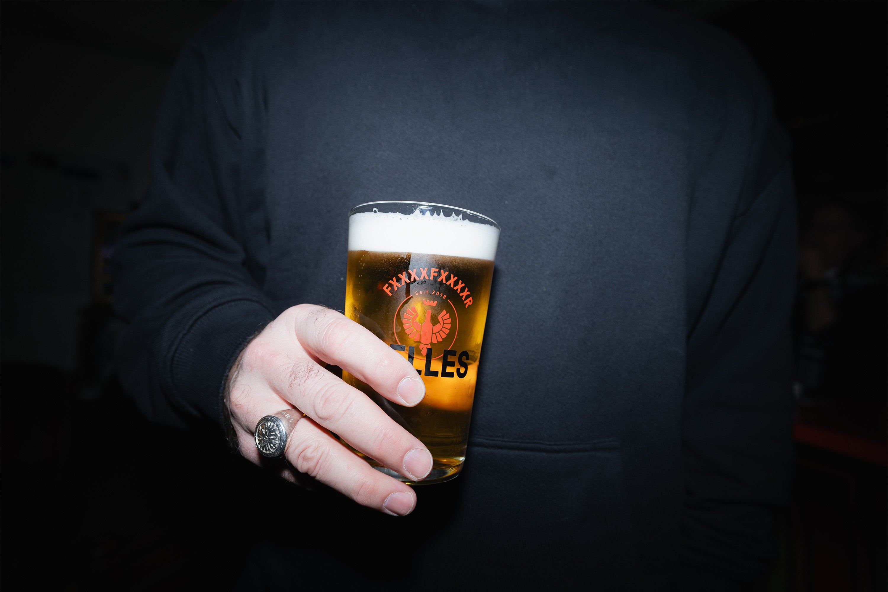Eine Person in einem dunklen Sweatshirt hält ein Bier mit schaumiger Schaumkrone und einem Logo auf dem Glas.