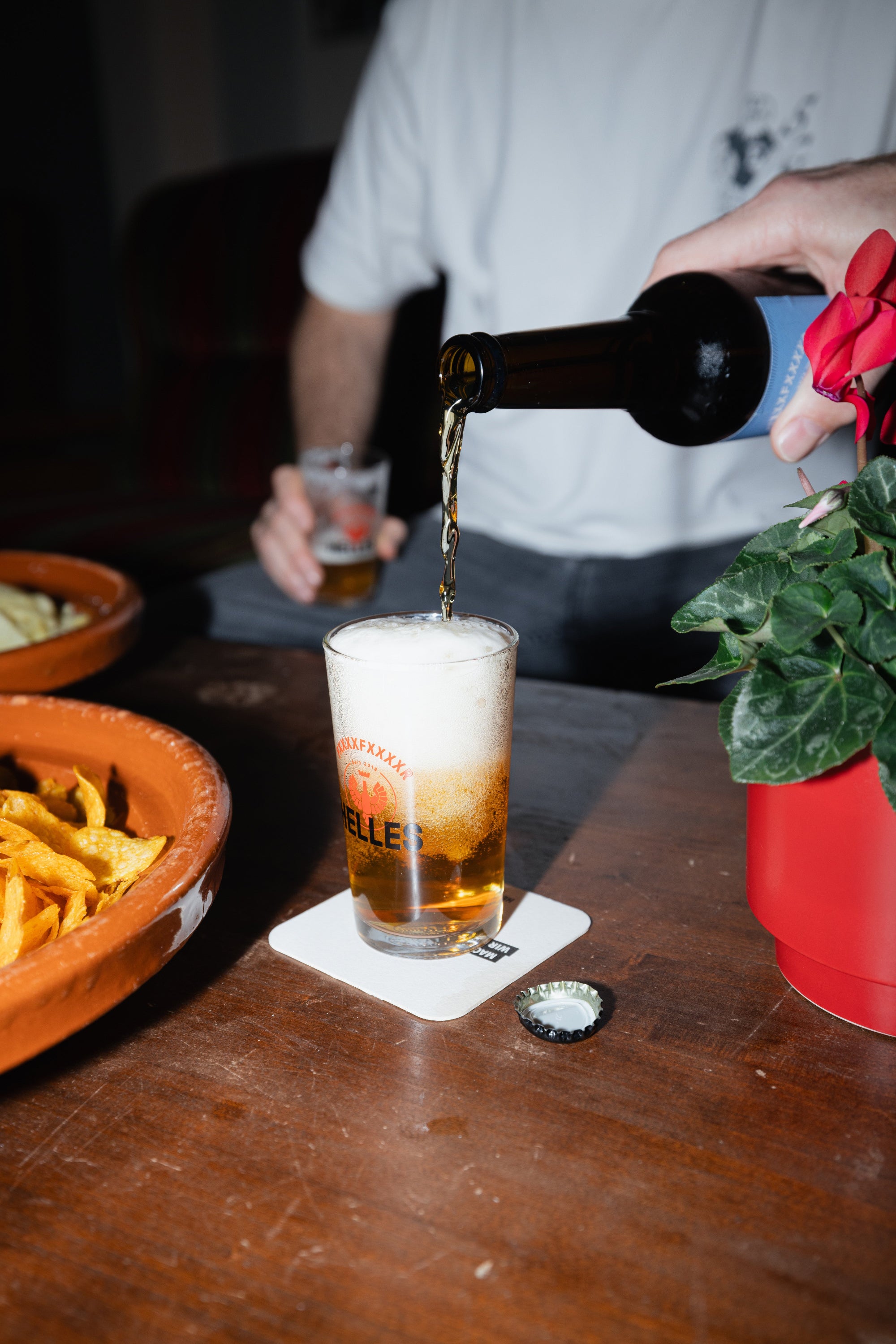 Eine Person gießt Bier in ein Glas auf einem Holztisch mit Chips, einem Untersetzer und einer Topfpflanze in der Nähe.